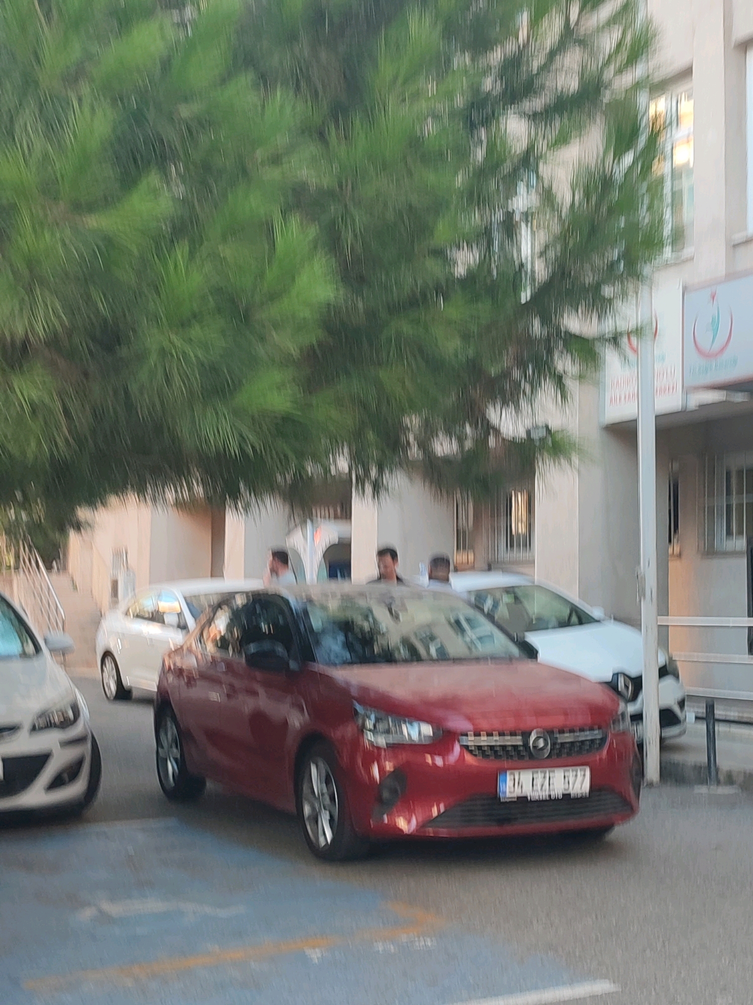 The image depicts a street scene in Kadıköy, Turkey, with a focus on several vehicles and a building. A red Opel Corsa is parked prominently in the foreground, displaying a license plate that reads "34 EZF 577". To its left, the front of a white car is partially visible. Behind the red car, a white Renault Clio is parked. The building in the background appears to be a healthcare facility, indicated by a sign with a red crescent and star emblem, a common symbol for Turkish healthcare services. Tall green pine trees with long needles are casting shadows and partially obscuring the view of the building and the street. Several individuals are discernible in the background, standing near the entrance of the building, but their features are indistinct due to the image's blurriness. The overall lighting suggests it is daytime.