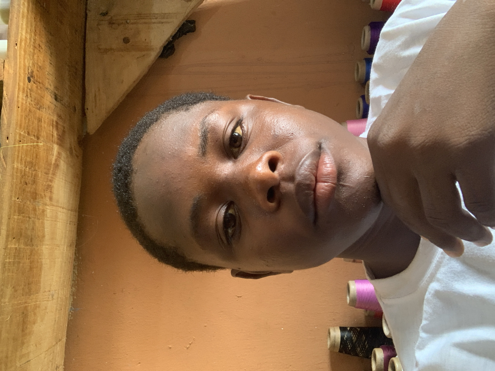 A young person, appearing to be male and of African descent, is shown in a close-up, head-and-shoulders portrait. He has dark skin and short, dark hair. The person is looking directly at the camera, with his chin resting on his right hand. He is wearing a white t-shirt with a crew neck.
The background consists of an orange-colored wall, likely painted. Above the wall is a wooden structure, possibly shelves or part of a ceiling, with visible grain and some darker markings. To the left and right of the person, in the background, are spools of thread in various colors, including black, pink, blue, and purple. Some of these spools are on what appears to be a shelf or ledge.
The lighting suggests an indoor setting, possibly during the day, with light sources casting soft shadows. The overall impression is of a casual, personal photograph taken in a domestic or workshop environment.
Based on the provided context that this image is located in Jos, Nigeria, it is plausible that the setting is a home or a small business, such as a tailor's shop or a craft studio, within the city. The presence of numerous spools of thread supports the idea of a place where sewing or textile work is done.