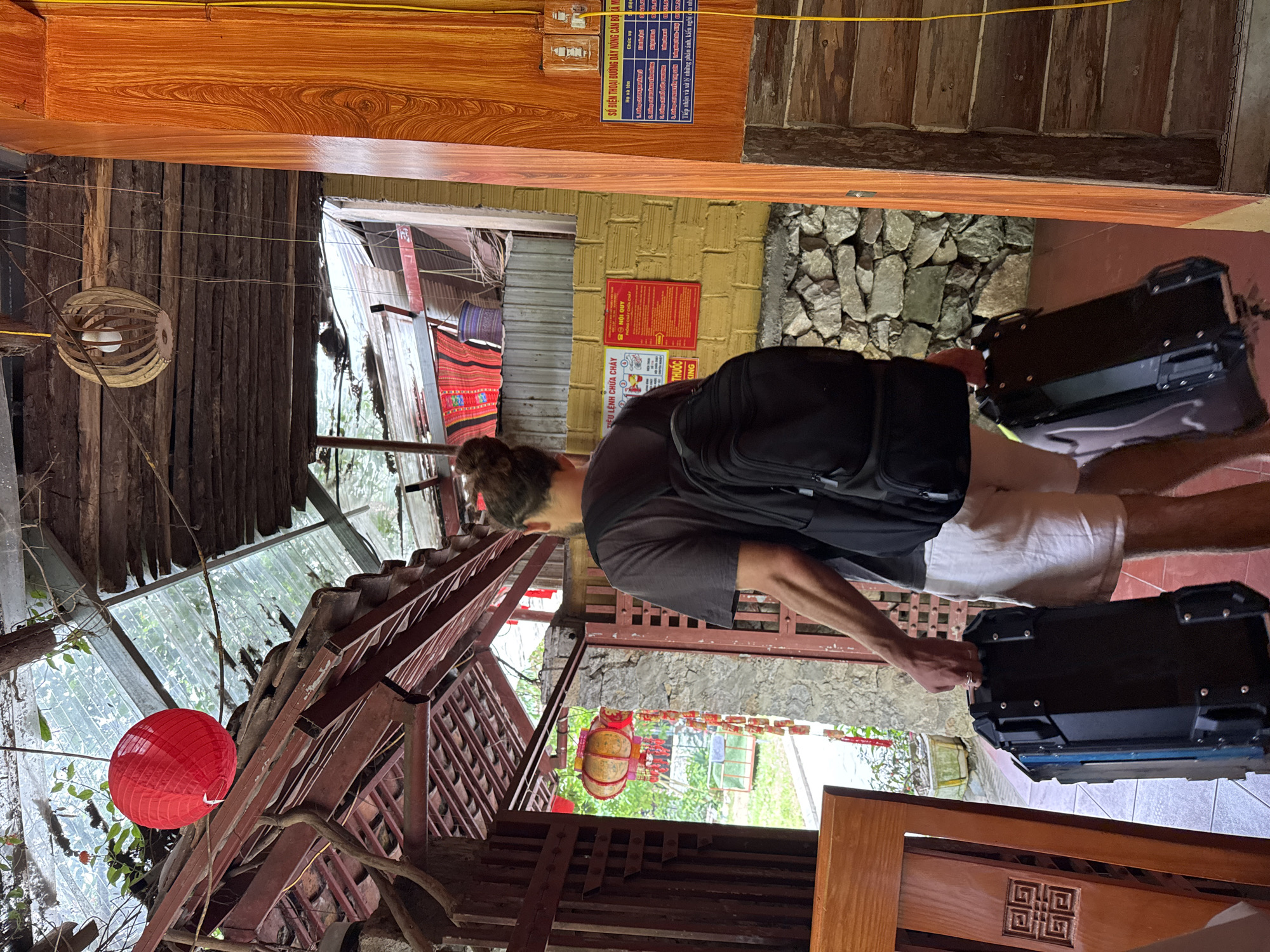 Here is a description of the image:
The image captures a person standing within a building in what is described as Mèo Vạc Commune, Vietnam. The individual, who is wearing a gray shirt and light-colored shorts, has a black backpack and is holding onto two black luggage cases with a blue trim.
The interior of the building features a mix of construction materials, including wood, stone, and brick. Decorations such as a red lantern and woven lamps are visible. Wall-mounted signs with Vietnamese text are present. The building's structure is partially open to the outside, showcasing foliage and other buildings in the background.