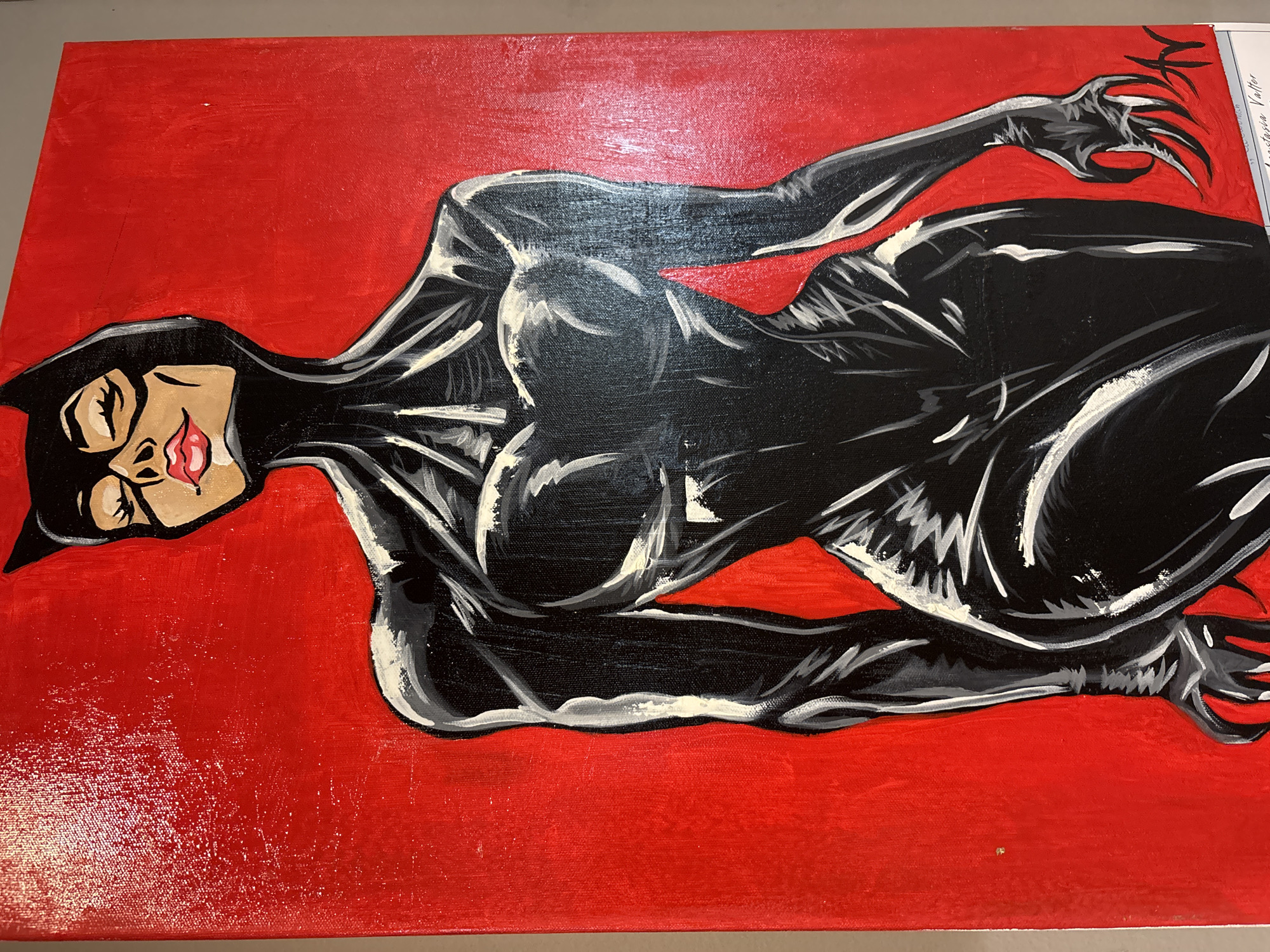 A horizontally oriented photograph depicts a painting of Catwoman on a red canvas. Catwoman, wearing a black catsuit and mask, is positioned in a reclining pose. Her skin is painted a tan color, and her lips are red. White and grey paint accents the black suit to create highlights. Her hands are flexed with extended claws. The background is painted a uniform shade of red. A piece of white paper is attached to the upper right corner of the canvas.