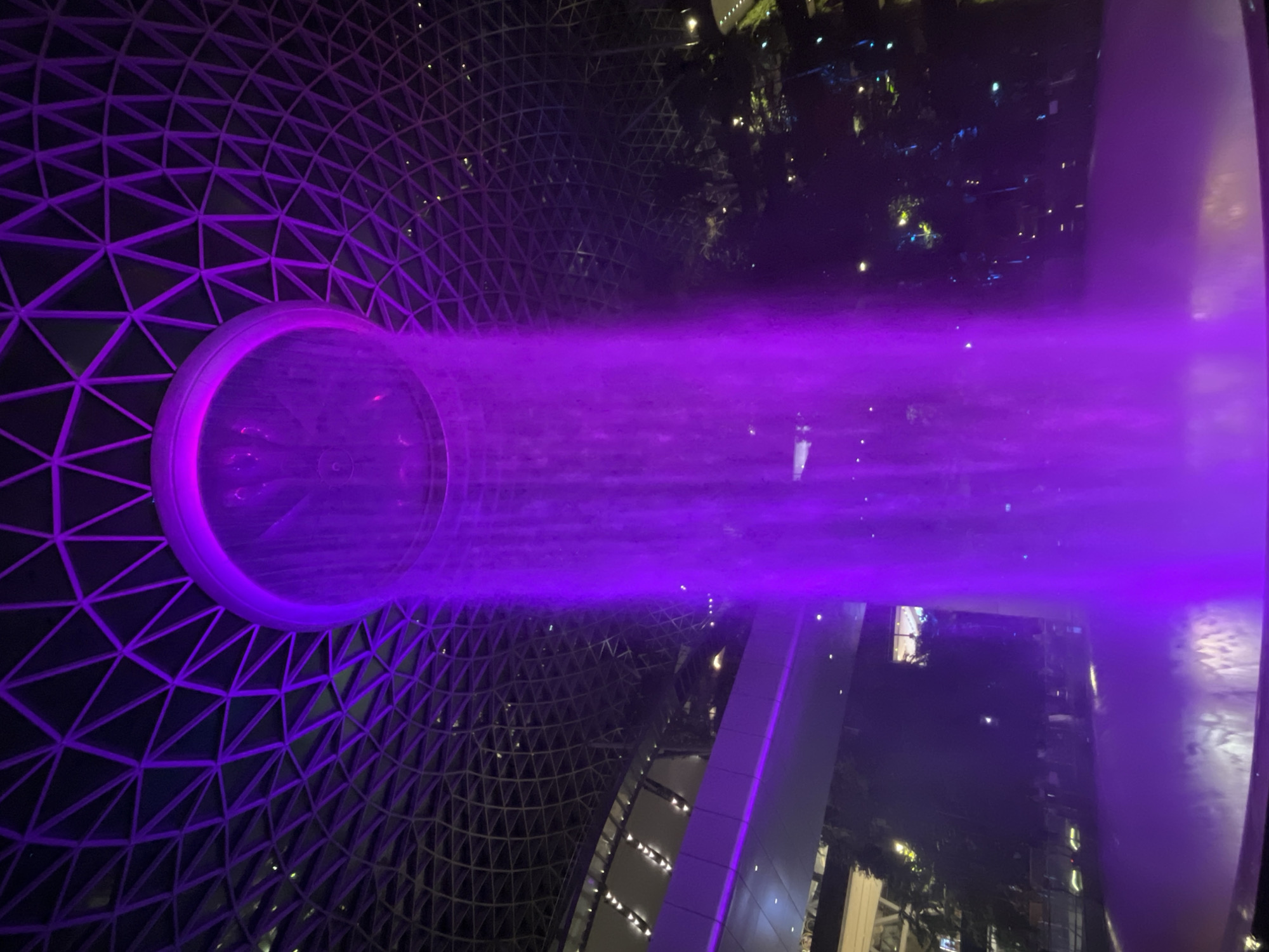 The image captures an indoor scene at night, likely within a modern architectural structure. The primary focus is a large, circular water feature emitting a vibrant purple mist or spray that extends horizontally across the frame. This water feature is set against a backdrop of a geodesic dome-like ceiling structure, characterized by a repeating triangular pattern, also illuminated with purple light.
In the background, beyond the misty curtain, glimpses of other levels of the building are visible. These appear to be indoor gardens or spaces with trees and artificial lighting, suggesting a commercial or entertainment complex. Some lights twinkle in the distance, adding to the nighttime ambiance. The overall atmosphere is futuristic and slightly dramatic, enhanced by the intense purple lighting.
There are no discernible people in the image. The scene appears to be an observation or public area designed to showcase the impressive water installation and the architectural design. The time of day is clearly night. The location is identified as Singapore. No text is visible in the image.