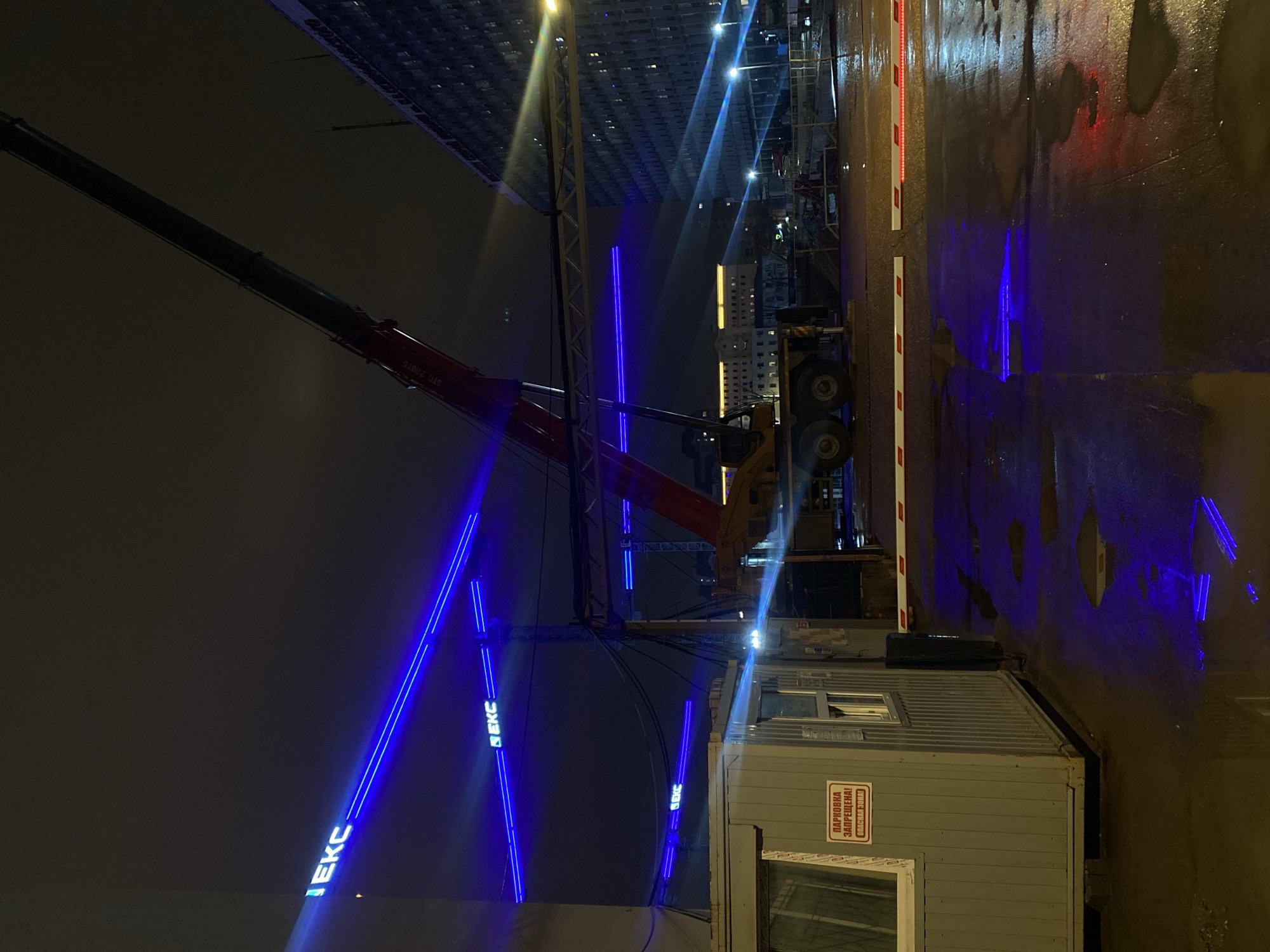 The image shows a night scene at a construction site in Moscow, Russia. A large crane with "EKC" written on it in blue lights stands near a modular construction office. The ground is wet, reflecting the blue lights and other ambient lights. The crane's boom extends diagonally across the frame, and the background shows other buildings and construction activity. A barrier with red and white stripes is also visible.