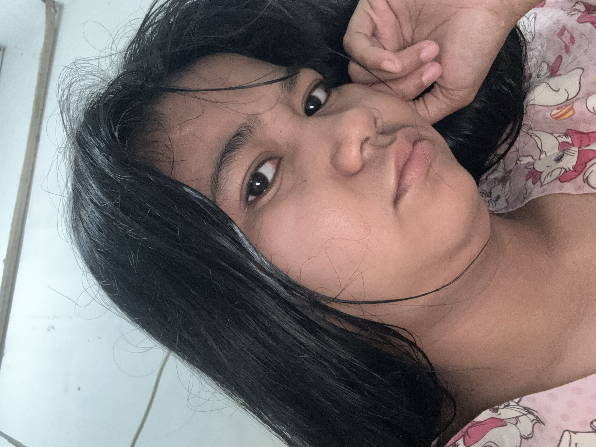 A close-up, head-and-shoulders selfie of a young woman with dark, straight hair is captured from a slightly high angle. Her head is tilted to the right, and her right hand is resting on her cheek, partially obscuring her face. Her hair is dark and appears slightly damp, with some strands falling across her face. She has a neutral expression, with her eyes looking directly at the camera. Her skin tone is light brown, and she is wearing a pink top with a white and pink cartoon pattern, possibly of cats.
The background is a plain, off-white wall with a horizontal wooden beam running across the top. The lighting suggests an indoor environment, possibly a bedroom or living space. There are no specific landmarks visible that would indicate the precise location within North Jakarta, Indonesia, beyond it being an indoor setting.