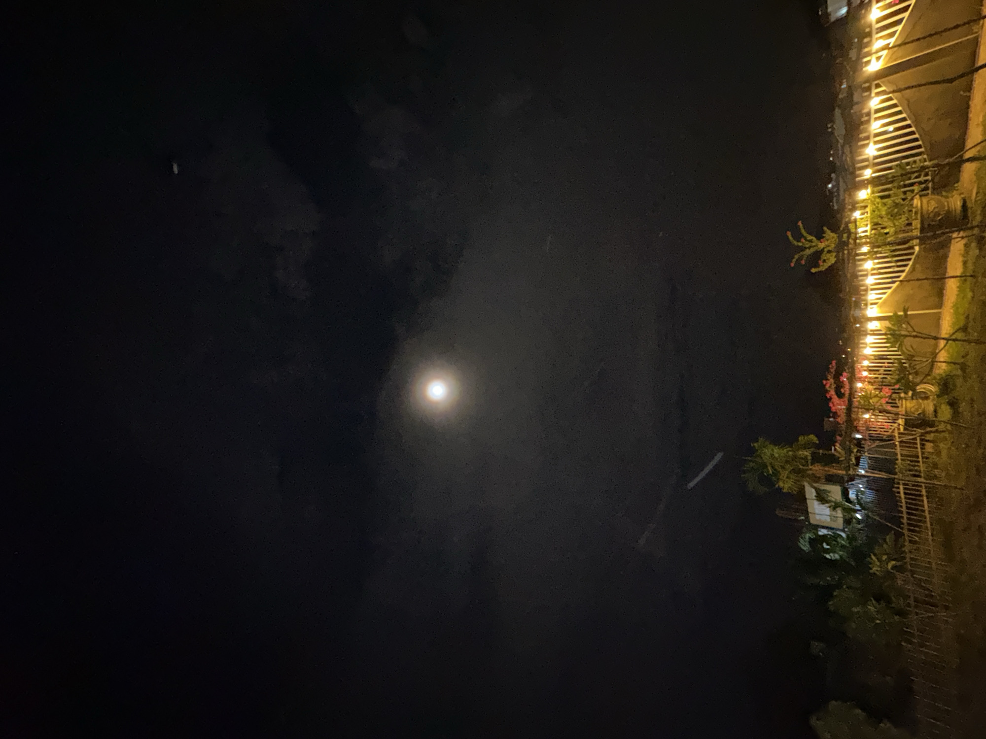The image shows a nighttime scene. On the right side, there is a structure with lights, possibly a fence or a building, and some trees. A bright light source, likely the moon, is visible in the middle of the sky. The location is in or near Bac Nha Trang Ward, Vietnam.