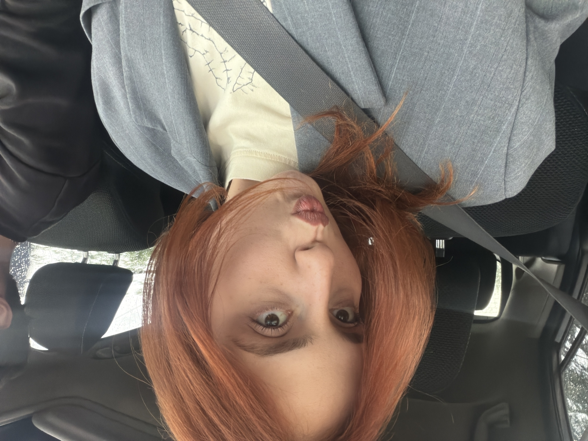 A woman with auburn hair is photographed from a close-up, upside-down perspective inside a vehicle, presumably a car. She is wearing a light-colored shirt with a design and a light grey blazer. A seatbelt is fastened across her. The interior of the vehicle includes a dark-colored seat and headrest visible in the background. The exterior of the vehicle, visible through the windows, appears to show wet conditions, with trees and sky visible. While a specific location within Tbilisi, Georgia, cannot be determined from the image alone, the presence of the vehicle indicates that the woman is likely traveling within the city. Another person is visible at the side, only partially.
