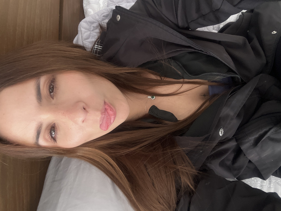 A young woman lies on a gray pillow. She sticks out her tongue slightly. Her hair is medium brown, and she has freckles on her nose. She wears a black puffy jacket. A silver necklace with a green pendant rests on her chest. The image is taken from an overhead perspective with the camera rotated.
