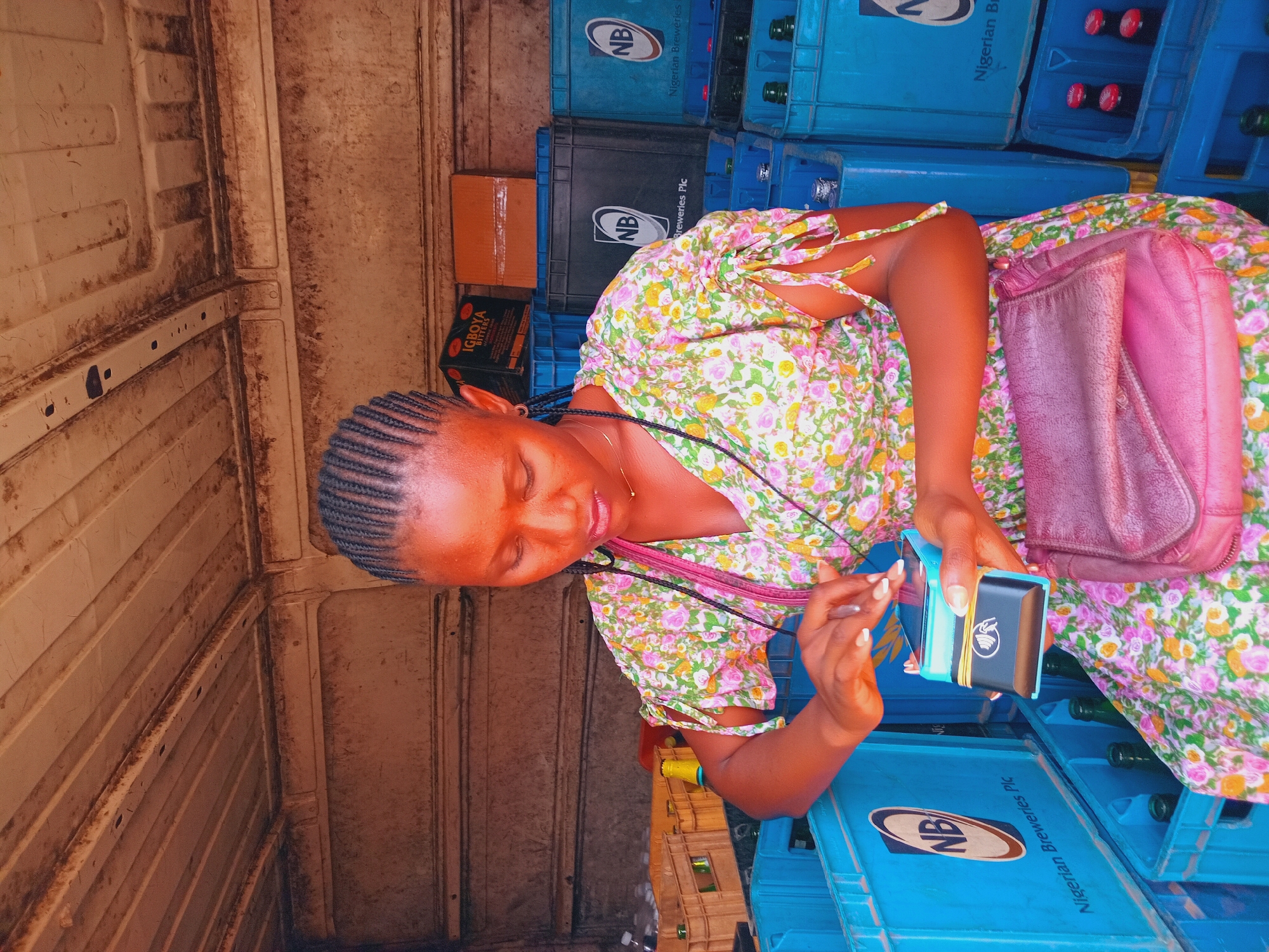 A woman sits in the back of a truck, surrounded by blue crates from Nigerian Breweries Plc. The crates contain beer bottles. She wears a floral dress and a pink purse hangs to her side. She is holding a blue wallet with a black stripe, containing money. The interior of the truck is lined with white panels. Based on the crates, this scene may be within Jos, Nigeria.