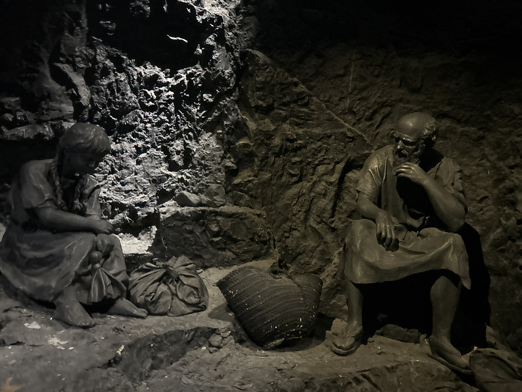 The image depicts a dark, rough-hewn cavern-like environment, possibly a mine, with two life-sized statues. On the left, a child statue, appearing to be a young girl with braided hair, is seated, looking down with a gathered cloth bag beside her. On the right, an elderly man statue is seated, his hand to his chin in a pensive pose. A woven basket sits between the two figures, and another gathered bag is near the girl. The statues are dark gray, appearing to be made of stone or a similar material, set against a similarly colored, textured background of rock. The lighting is dim, casting deep shadows, enhancing the somber atmosphere. This scene is located within the Salt Caves in the city of Tamar Regional Council, Israel.