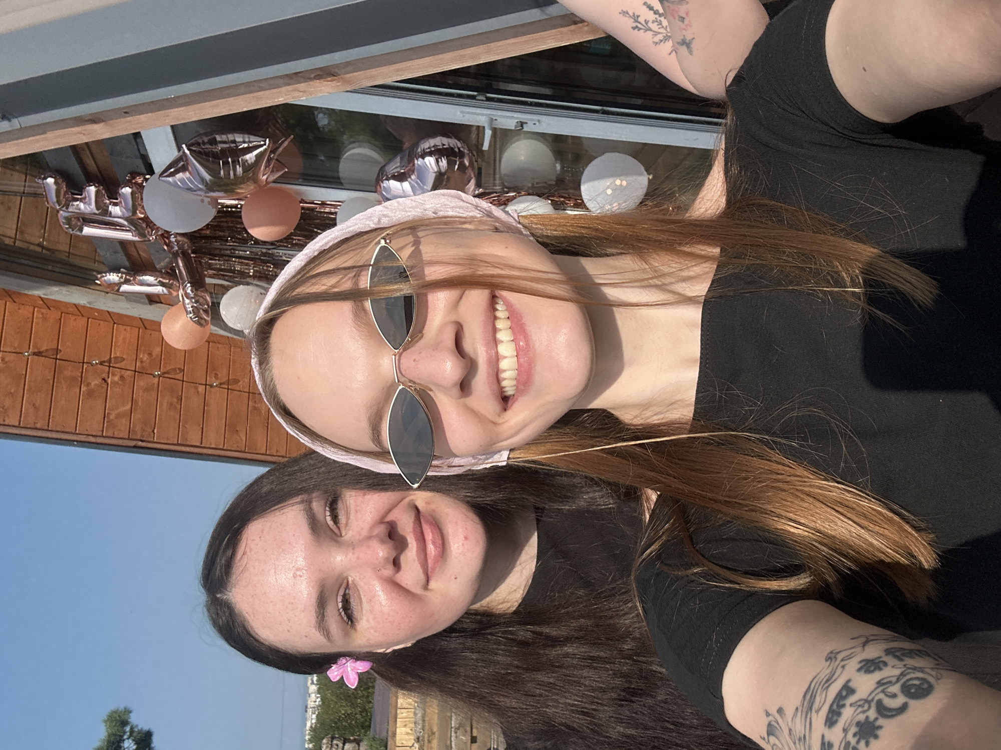 Two young women are posing for a selfie outdoors during the day. The woman in the foreground is on the right, wearing sunglasses and a pink headscarf. She has long auburn hair and is smiling with her teeth showing. She has a tattoo on her arm. The woman behind her is on the left, with long dark hair and a pink flower tucked behind her ear. She is also smiling. They are both wearing black shirts. In the background, there are balloons and a wooden structure, possibly a house or gazebo. The sky is clear and blue. The environment suggests a celebratory or casual outdoor gathering.