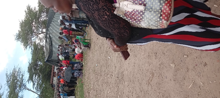 The image depicts a gathering of people in Kitengela, Kenya, at coordinates -1.47 latitude and 36.959 longitude. The scene is set in an open area with a dirt ground. A large group of people are standing and sitting under a partially collapsed corrugated iron shed. Several trees are visible in the background, while the foreground features a woman wearing a black lace top, a striped red and white skirt and carrying a polka-dot handbag, standing with her back turned towards the camera, her hands holding a wooden object, presumably a walking stick. The woman is looking at the people gathered in the distance. The scene suggests a public event or gathering in the rural area, perhaps a social gathering, meeting, or religious event.