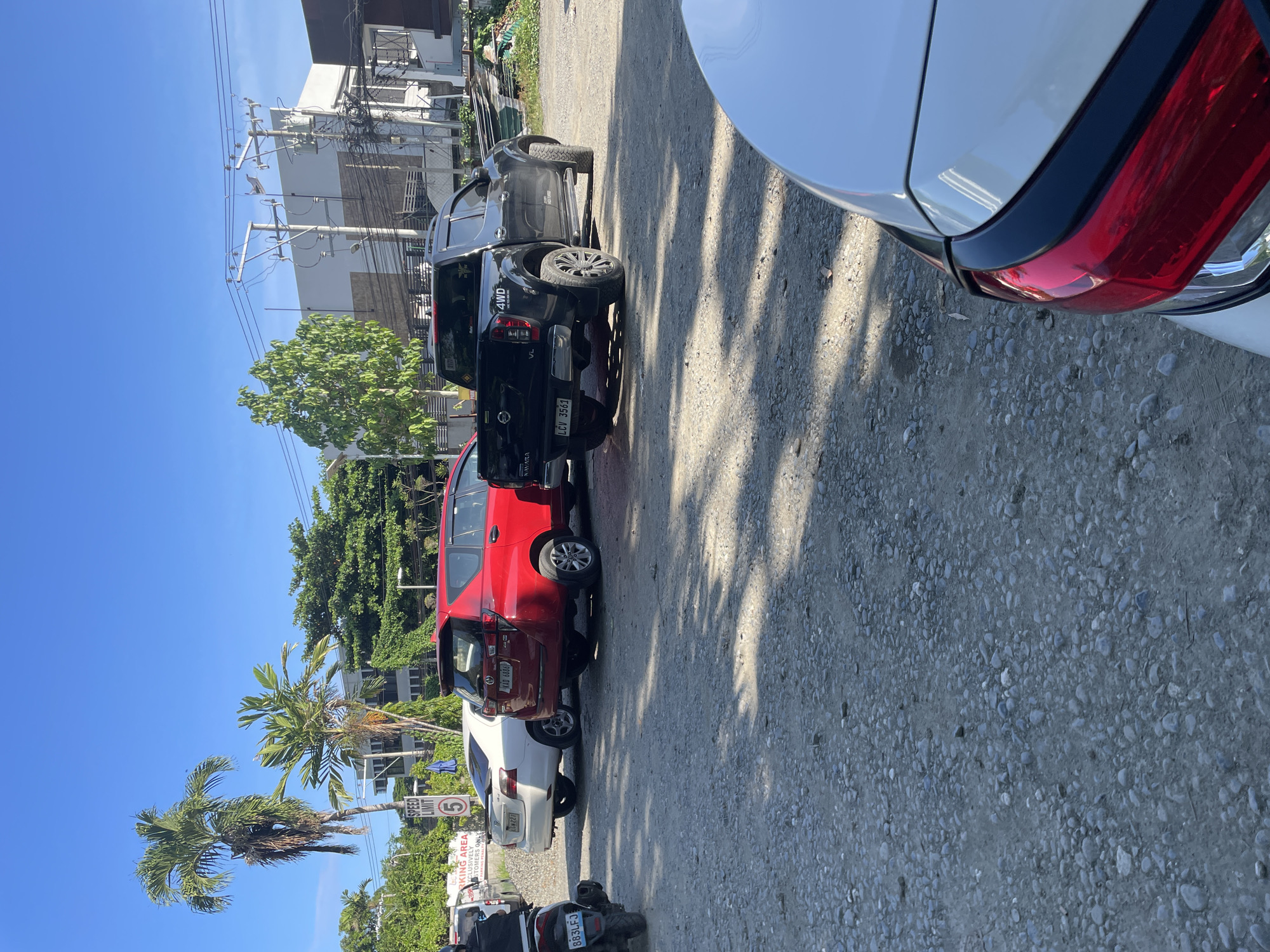 This image captures an outdoor, unpaved parking area under a clear blue sky in Davao City, Philippines. The ground is a mix of gravel and dirt, with elongated shadows cast by vehicles and trees, indicating direct sunlight, likely morning or late afternoon.
In the midground, from left to right, are several parked vehicles. A white Toyota Vios sedan (license plate CJK227) is parked, facing away from the viewer. To its right is a red Toyota Innova minivan/SUV (license plate KAB 6888), also facing away. Further right, a black Nissan Navara pickup truck (license plate LCV 3561) is parked, also facing away. On the far left, partially visible, is the rear section of a motorcycle, possibly a scooter, displaying the license plate 883LFJ. In the immediate foreground, the white rear bumper and red tail light of another vehicle are partially visible.
Various trees are present in the scene, including tall palm trees on the left and several broadleaf trees. In the background, multiple multi-story buildings with white and brown facades are visible, characteristic of urban architecture. A network of utility poles and overhead power lines spans across the background. Two signs are visible on the left: a rectangular sign, partially obscured, which reads "PARKING AREA EXCLUSIVELY FOR CUSTOMERS ONLY," and a circular "SPEED LIMIT 5" sign. No people are discernible in the frame.