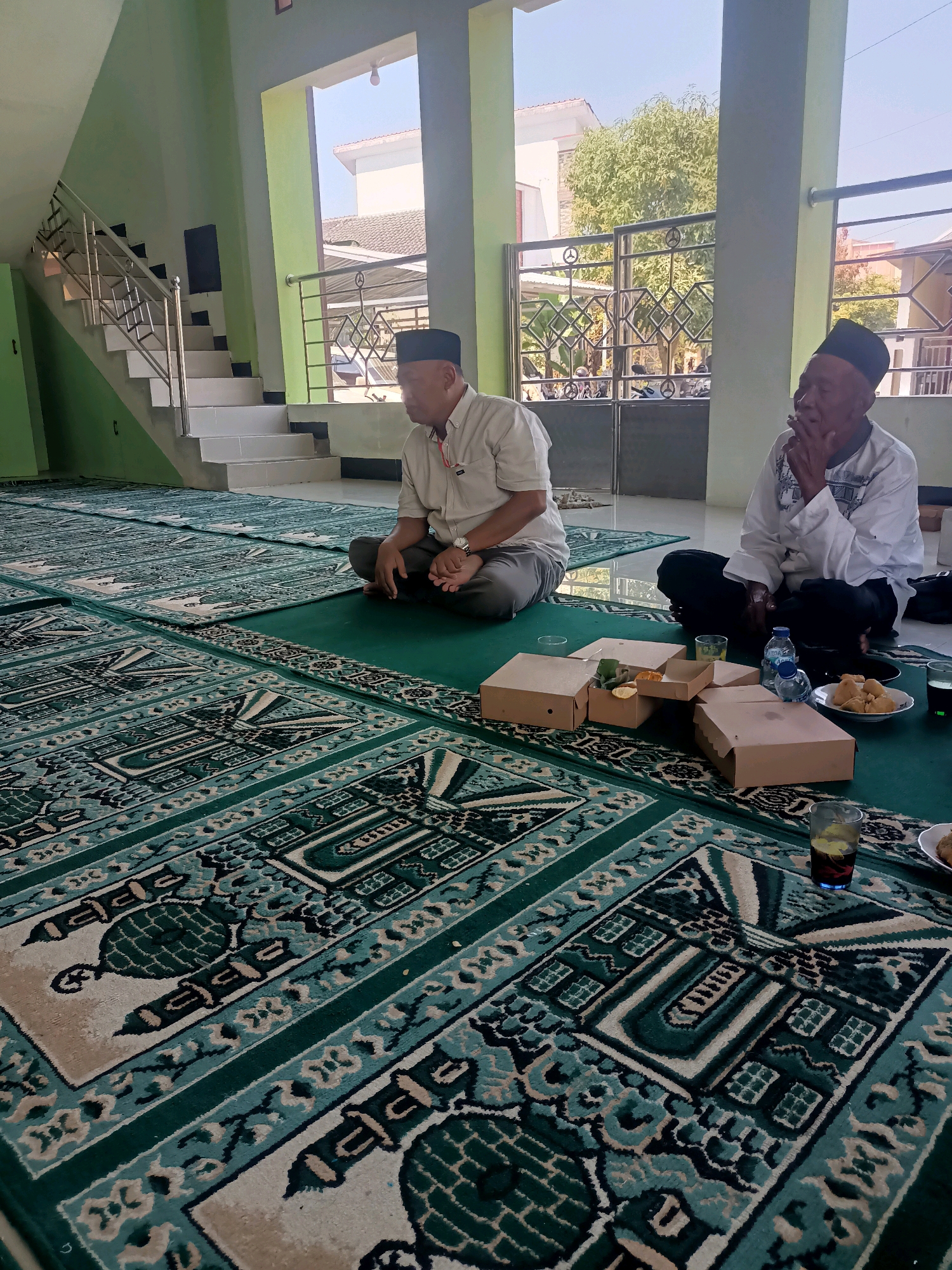 Two elderly Indonesian men sit on prayer mats in a hall. The man on the left wears a light-colored collared shirt and dark pants, with a black cap. The man on the right wears a white long-sleeved shirt with blue accents and a black cap, and he is smoking. In front of them are several cardboard food containers, some open, with food and fruit visible, a plate of fried snacks, glasses of drinks, and a bottle of water. The room has light green walls, a staircase with a metal railing to the left, and large windows or open doorways revealing a sunny outdoor scene with trees and buildings. The prayer mats have a repeating pattern in green and cream. The scene appears to be in a public building, possibly a mosque or community hall, in Penusupan, Indonesia.