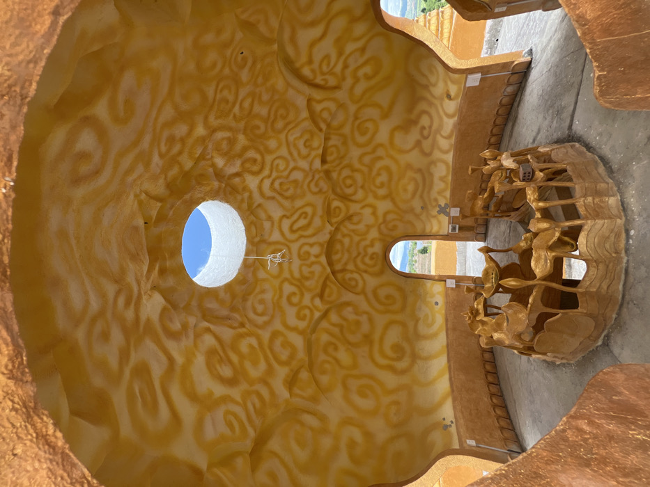 The interior of a circular, domed structure in Đăk Mar, Vietnam, is captured from a low-angle perspective. The ceiling and upper walls are a textured, molded surface painted in an earthy orange-brown base with a lighter yellow-orange swirling, cloud-like pattern. A circular oculus at the dome's apex reveals a clear blue sky, framed by a prominent white cylindrical lip. A white, knotted string hangs from the center of the oculus. The lower walls are a darker brown, featuring a simple base molding. The floor is a flat, light gray concrete surface. Centrally located on the floor is a large, circular, sculpted structure painted in a uniform golden-brown. This central piece features multiple organic, stylized elements resembling lotus flowers, leaves, and tiered basins, configured as a dry fountain or altar. Two arched doorways are visible in the far wall, opening to a brightly lit exterior landscape of green vegetation and distant hills under a blue sky. No individuals, actions, or interactions are present within the scene.