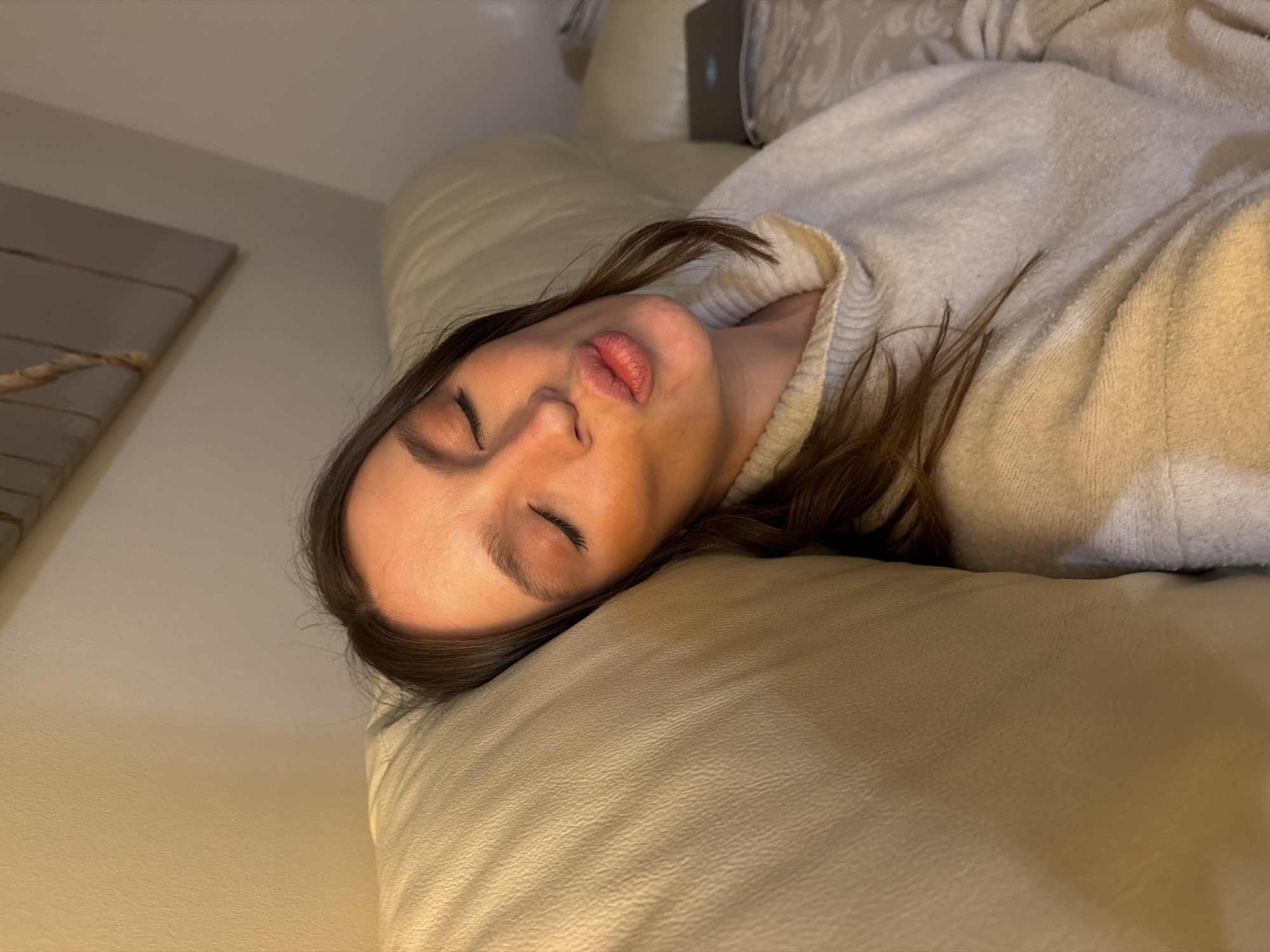 A female individual is depicted indoors, situated in Budva, Montenegro. She is positioned against the back cushion of a light beige, textured sofa, with her head resting on the cushion. Her eyes are closed, and her lips are pursed into a distinct kissing or puckered expression. Her long, dark brown hair flows around her shoulders. She is dressed in a light-colored, likely cream or beige, knitted turtleneck sweater. The background consists of a plain, light-colored wall. Above the sofa, a horizontal piece of wall art featuring a twisted rope design is mounted. To the individual's right, a closed, dark-colored laptop is visible resting on a patterned surface, possibly a blanket or another sofa cushion. The scene is illuminated by warm, artificial light.
