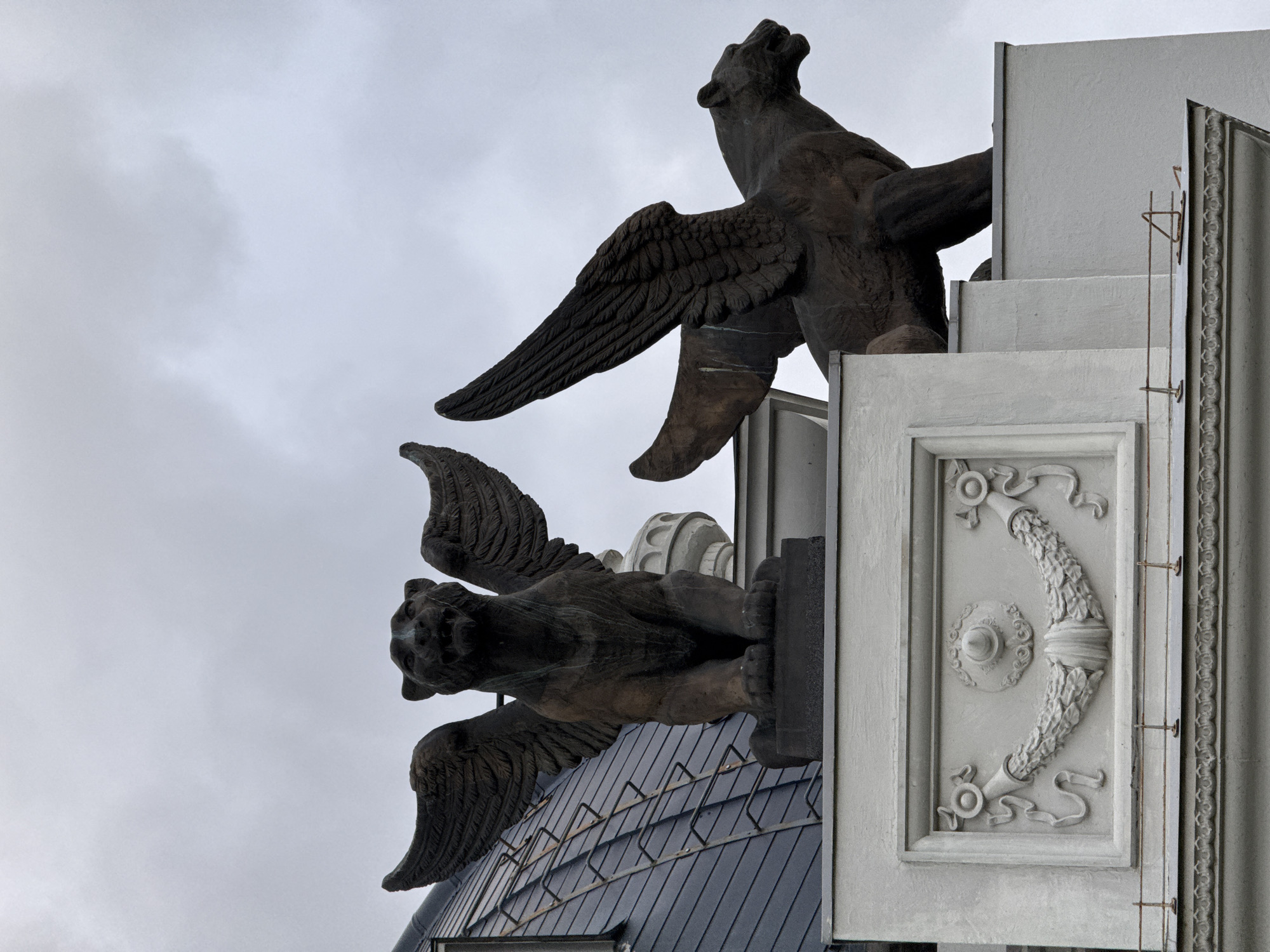 The image depicts two large, dark bronze sculptures of winged mythical creatures, resembling griffins or winged lions, perched on the rooftop of a building in Kazan, Russia. The sculpture on the left is facing forward, with its wings spread, and is positioned on a dark pedestal. The sculpture on the right is seen in profile, with its wings also spread, and appears to be slightly lower than the one on the left. Both creatures have feline features with prominent wings.
The building itself features architectural details, including a dark, mansard-style roof with visible tiling and a ladder or railing system. Below the sculptures, the building's facade is adorned with a white, decorative element featuring a relief of a wreath with ribbons and a central medallion. The sky is overcast and grey, suggesting a cloudy day. The overall scene conveys a sense of grandeur and historical architectural style.