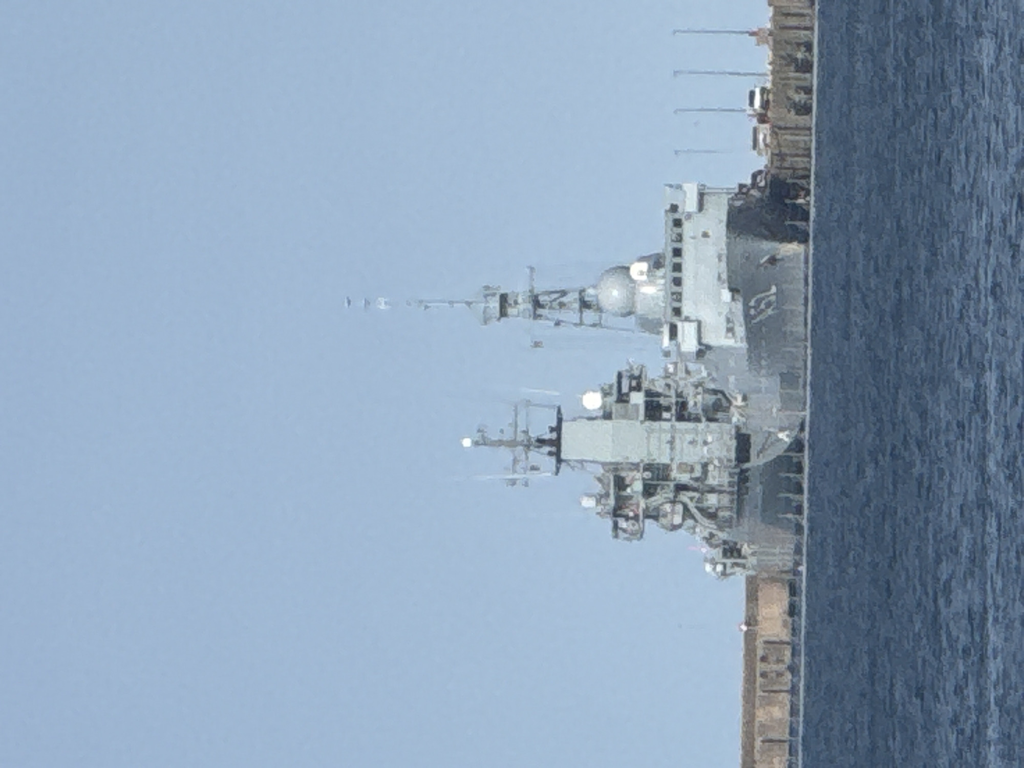 The image shows a large grey naval vessel docked at a pier on a clear day. The ship has multiple levels and appears to be a warship, with various antennas and structures visible. The water in the foreground is dark blue and choppy. Behind the ship, a wooden pier stretches along the shoreline, with several vehicles, including a white van, parked on it. The background is a clear, pale blue sky. The location is the Baltic Sea in Eckernförde, Germany.