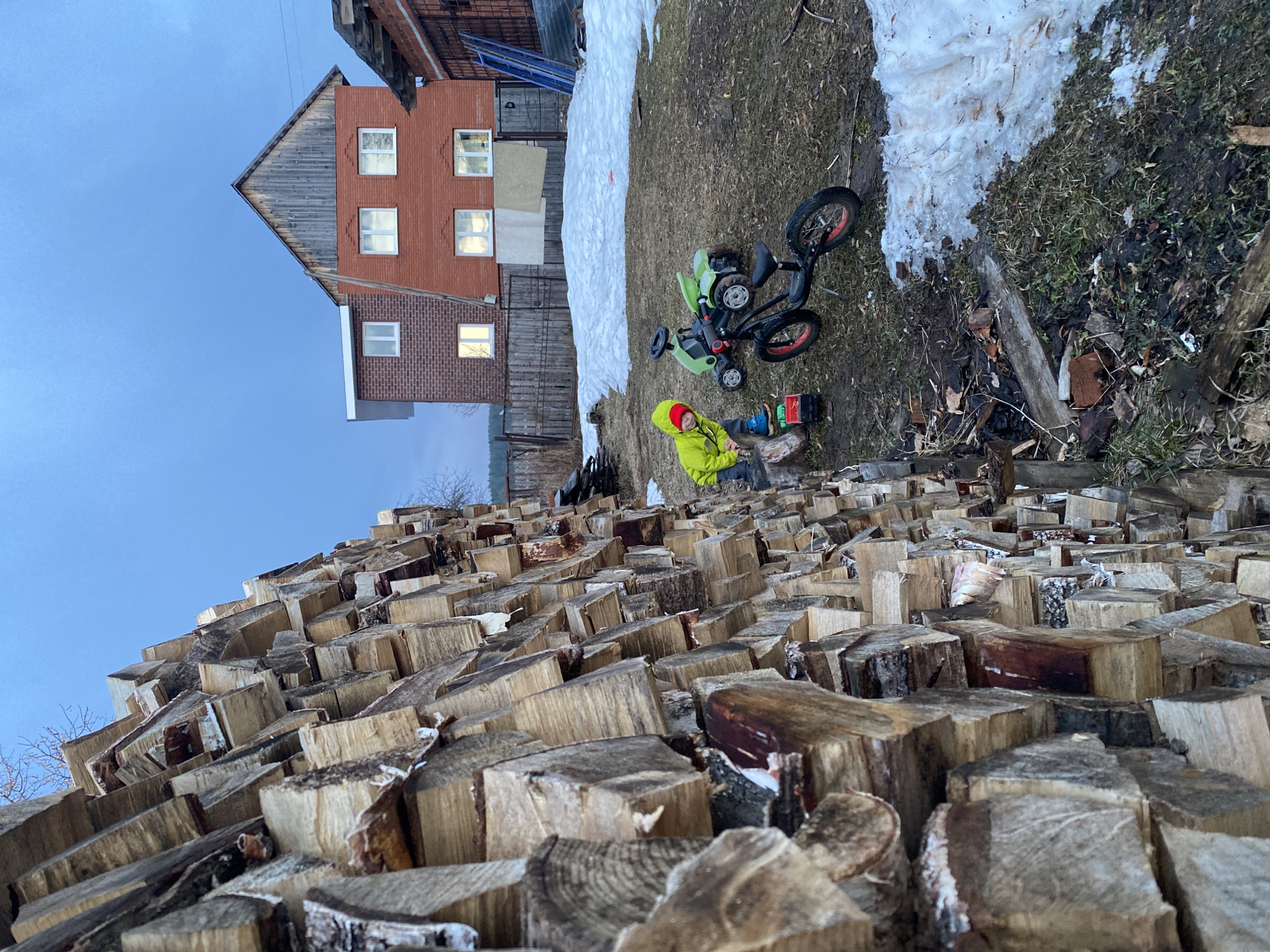 The image depicts a scene in Ольховка, Russia, with a large pile of logs in the foreground occupying the majority of the frame. A child in a yellow jacket and red hat sits among the logs. In the background, there is a patch of melting snow, a child's green toy car, and a residential building with lit windows. The sky is visible in the upper portion of the frame, indicating either dawn or dusk.
