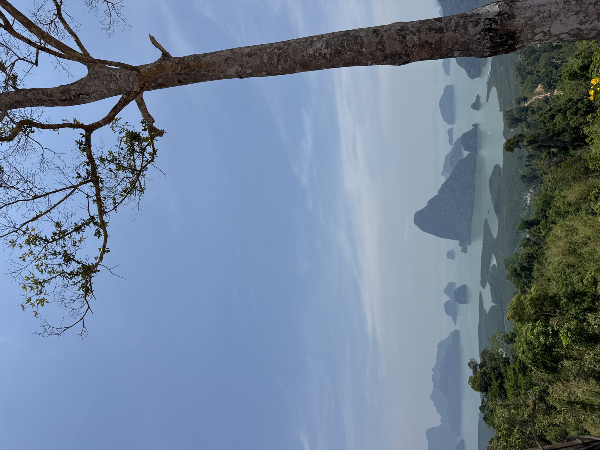 The image shows an elevated view in Ban Khlong Khian, Thailand. The lower right quadrant features dense green vegetation, suggesting a forested or hilly area. A body of water with several rocky islands or formations is visible in the middle, receding into a hazy background. A large tree branch extends across the top of the image, with smaller branches and sparse leaves appearing in the upper left. The sky is blue with thin, wispy clouds.