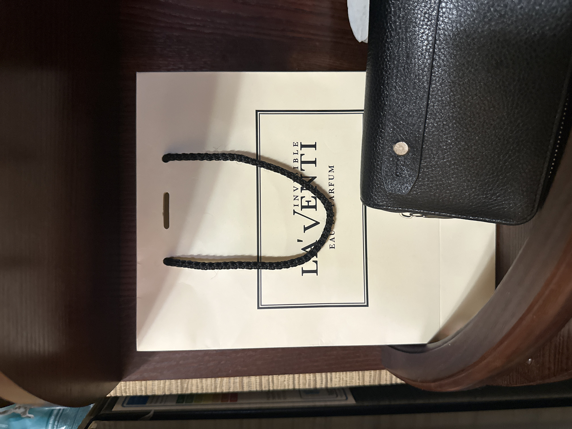 A light-colored shopping bag with black braided handles sits on a wooden shelf. The bag features a black-bordered rectangular logo with text that reads "LA'VENTI INVISIBLE EAU DE PARFUM". In front of the bag, a black textured wallet with a gold button closure is visible. The background is a dark wooden surface. The image appears to be taken indoors, possibly in a home or retail setting in Aleksandrovka, Russia.