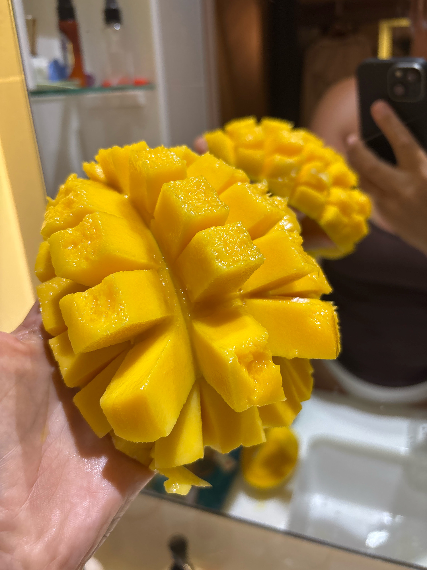 A person's hand holds a halved, star-shaped mango, cut into cubes. The mango is a vibrant yellow-orange color. In the background, a mirror reflects a bathroom sink, a phone, and another cut mango. The location is a bathroom, likely in Karon, Thailand, given the prompt's instruction.
