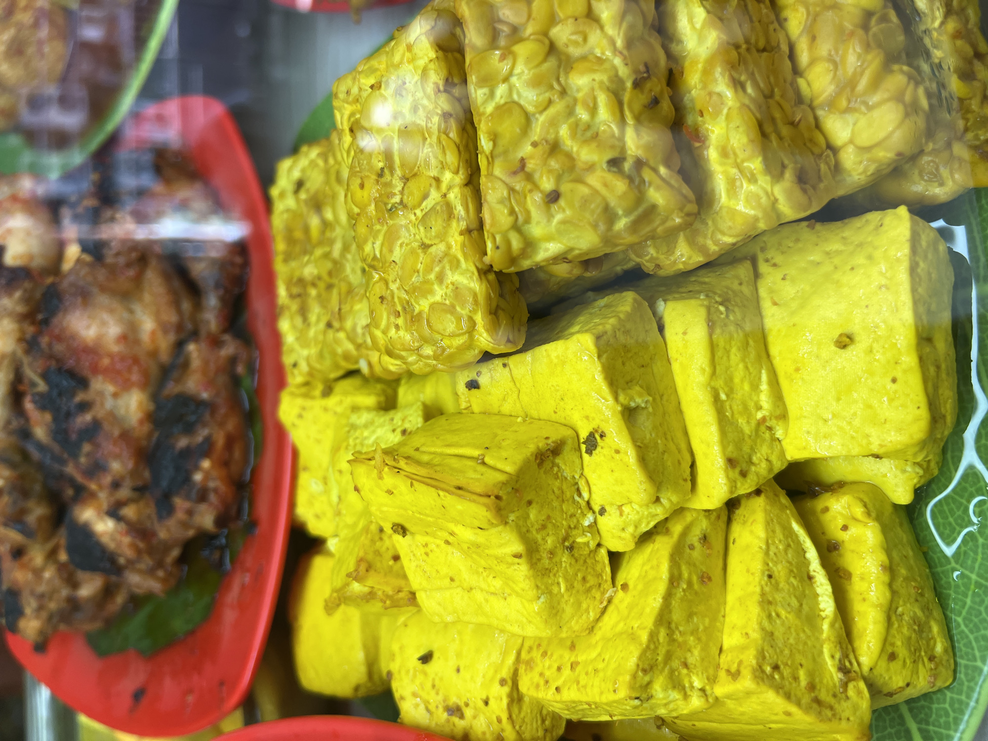 In Kuta, Indonesia, a close-up shot showcases a display of Indonesian food items. In the foreground, stacked blocks of bright yellow marinated tofu are prominent, some with visible spices embedded within. Adjacent to the tofu are pieces of yellow-marinated tempeh, with its characteristic bean texture. In the background, out of focus, a red plate holds grilled chicken pieces, also appearing to be marinated or seasoned. The overall setting suggests a food stall or market.