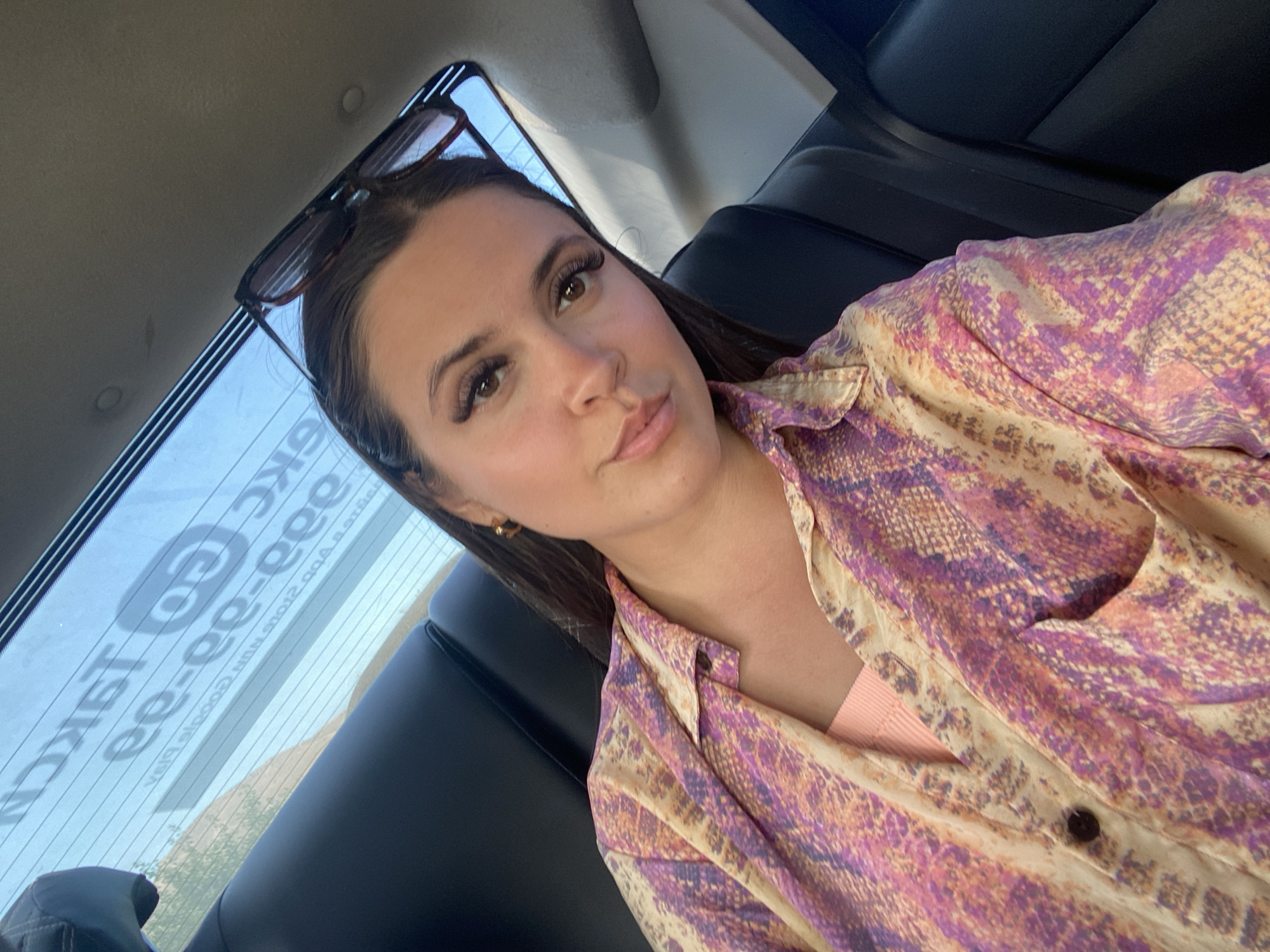 A young woman is seen from the chest up, taking a selfie inside a vehicle. She is wearing a long-sleeved, collared shirt with a snakeskin-like pattern in shades of pink, purple, and yellow. She has on a peach-colored tank top underneath. Her hair is dark brown and pulled back, with sunglasses perched on top of her head. The background shows the interior of a car, with a black leather seat visible on the right side and the rear window of the car on the left. The rear window has text on it, which is partially obscured, but appears to be an advertisement or logo. The scene is lit by natural light. There is no indication of the location being in Gelinbatan, Russia.