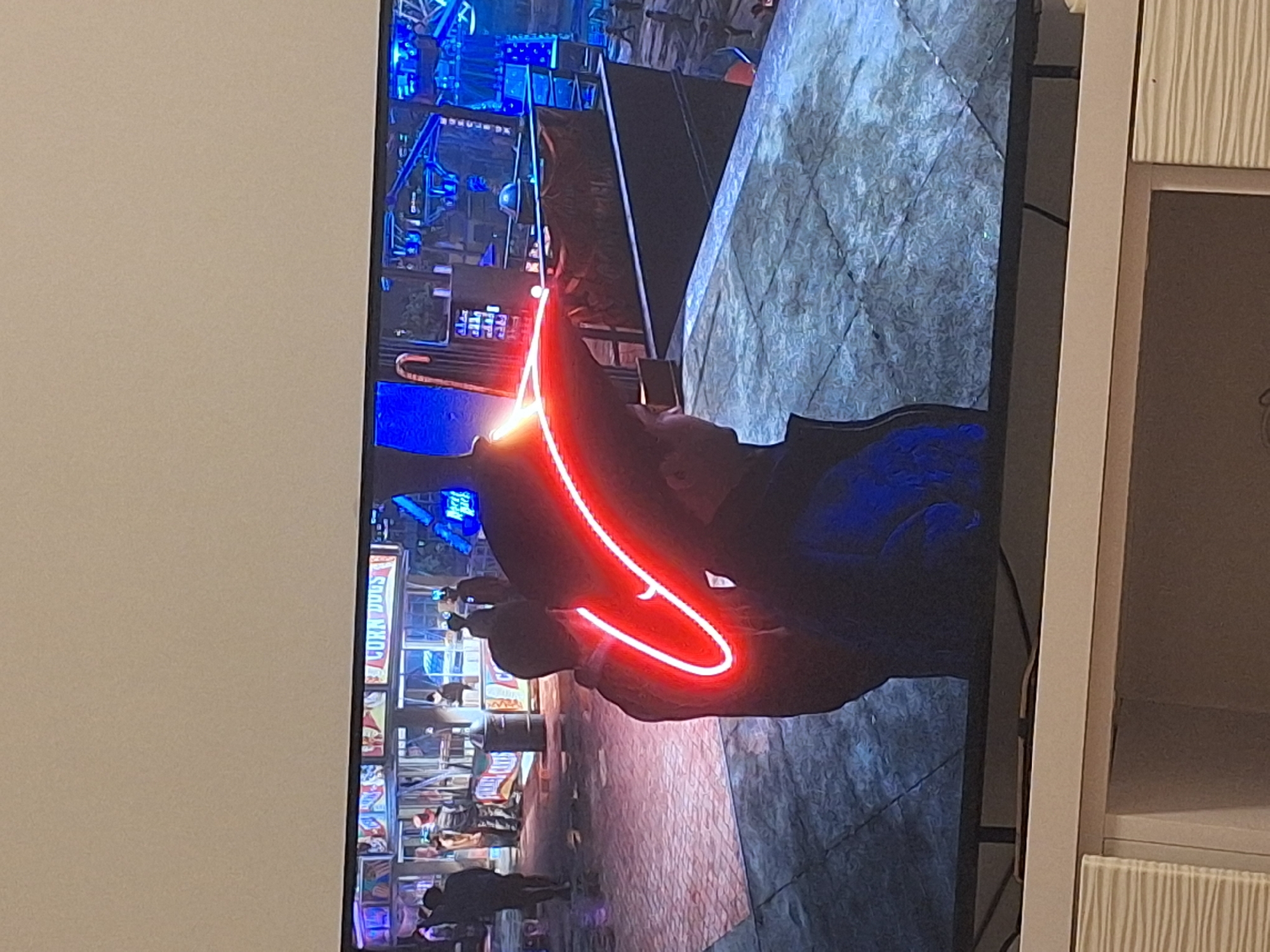 The image shows a television screen displaying a scene from a video game. The game scene depicts an outdoor, possibly nighttime setting with a concession stand labeled "CORN DOGS" in the background. Several indistinct figures, presumably game characters, are visible in the background, some near the stand and others walking. In the foreground, two game characters are the primary focus. One character is wearing a dark jacket and a hat with a bright red neon outline. The other character, to the left, is mostly obscured by shadow but appears to be wearing dark clothing. The environment is paved with a textured surface that resembles cobblestones or large tiles. The lighting in the game scene is dramatic, with prominent blue and red neon lights illuminating parts of the environment. There is no identifiable information in the image that relates to Mińsk Mazowiecki, Poland.