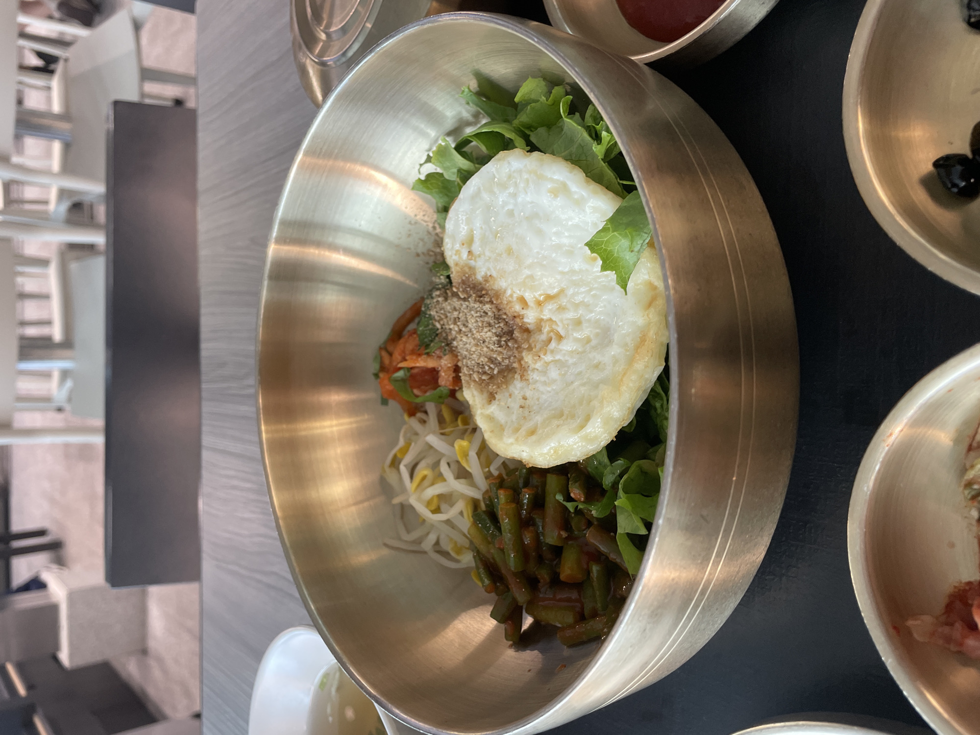 A close-up, eye-level shot captures a traditional Korean bibimbap served in a large, metallic brass bowl. The dish is artfully arranged with various ingredients, including vibrant green leafy vegetables, stir-fried green beans with a reddish sauce, pale yellow bean sprouts, and a fried egg with a runny yolk, all topped with a sprinkle of sesame seeds. To the left of the main bowl, a smaller white bowl contains a light-colored broth, possibly soup. The setting appears to be an indoor restaurant, with blurred background elements suggesting tables and chairs, and a hint of natural light. The table surface is a light-colored wood grain. The overall scene conveys a sense of freshly prepared, wholesome Korean cuisine, likely in a dining establishment in Incheon, South Korea.