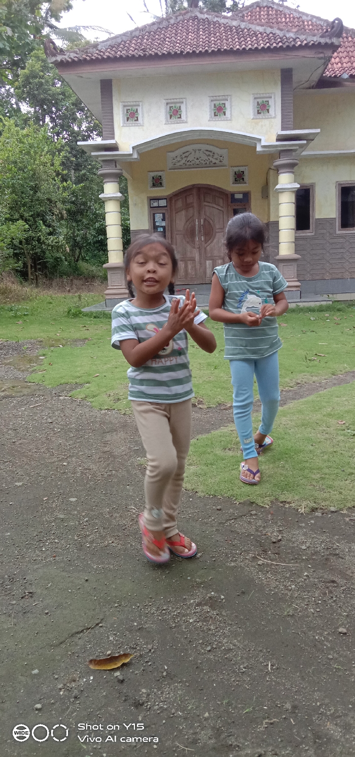 Two young Asian females are observed outdoors in Ranugedang, Indonesia, in front of a light yellow/beige building with a terracotta-tiled roof. The building features a double wooden door entrance, decorative trim, and square ornamental panels with red and green patterns. Two ornate support columns flank the entrance area.
In the foreground, a female child, approximately 5-7 years old, is in motion, with her left foot slightly raised. She wears a horizontally striped white and light green t-shirt with a bunny graphic and "HAPPY" text, light brown leggings, and pink/red flip-flops. Her dark hair is styled in pigtails. Her mouth is open, and her hands are held together in front of her chest, as if clapping or speaking.
To her right, a second female child, approximately 5-8 years old, stands on a patch of green grass. She wears a horizontally striped light green and white t-shirt, light blue leggings, and purple flip-flops. She is looking downward at her hands, which are held near her waist, appearing to examine a small object.
The immediate ground consists of dark gray gravelly dirt with sparse green growth, and a single brown leaf is visible on the ground. Beyond the grass, dense green foliage and trees are visible in the background.