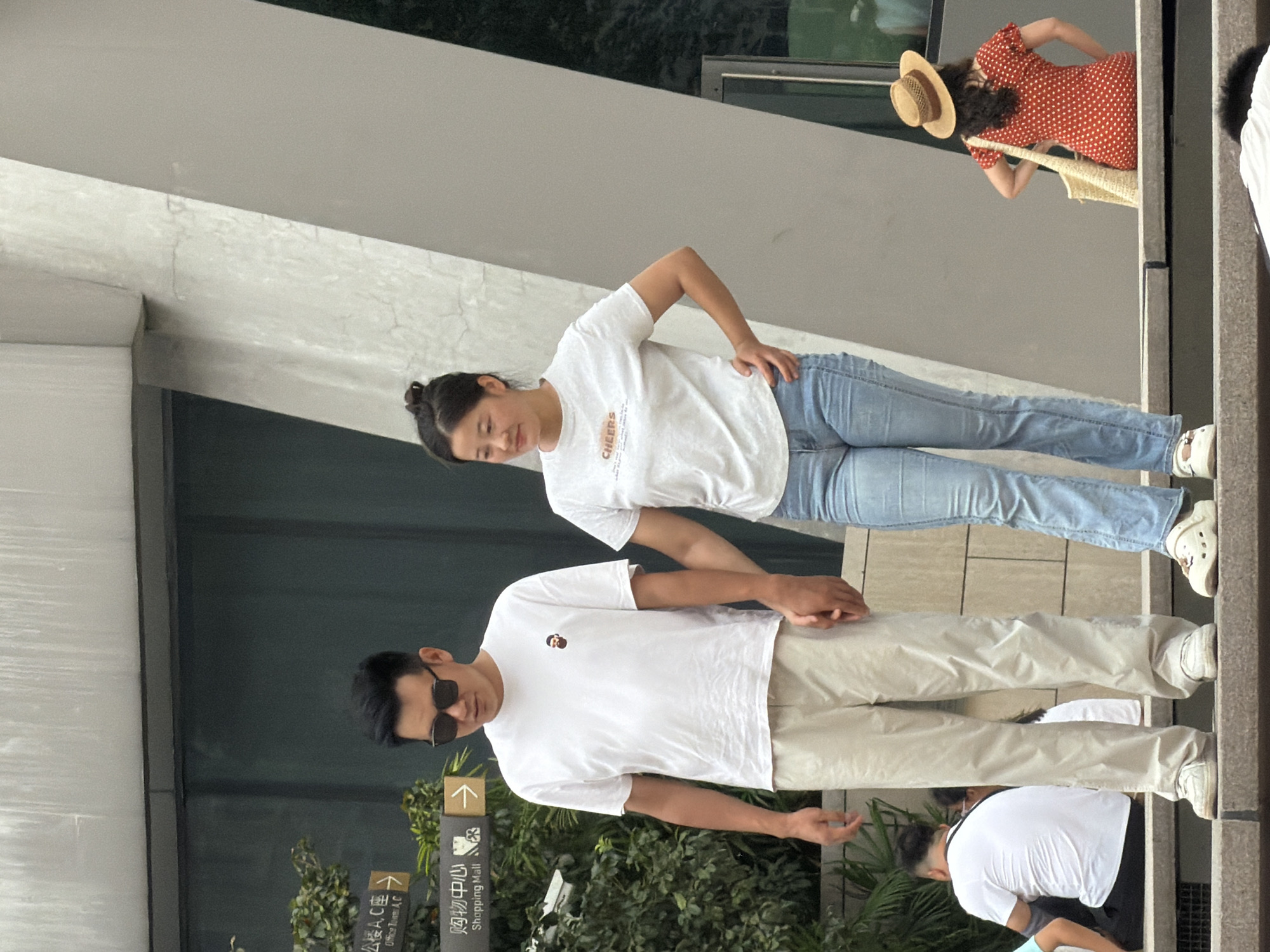 In Yuzhong District, China, a couple stands on a tiered stone structure outdoors. The man, on the left, wears sunglasses, a white t-shirt with a small cartoon image, and light-colored pants. He holds the hand of the woman next to him. The woman, on the right, wears a white t-shirt with the word "CHEERS" and light blue jeans. She has her left hand on her hip. In the background, to the left, are green bushes and directional signs pointing to "Office Tower A,C," and "Shopping Mall." To the right, a woman in a red polka dot dress and a straw hat sits with a straw bag. Other individuals are visible in the lower portion of the frame, sitting and facing away from the camera. The environment appears to be a public urban space with modern architecture.