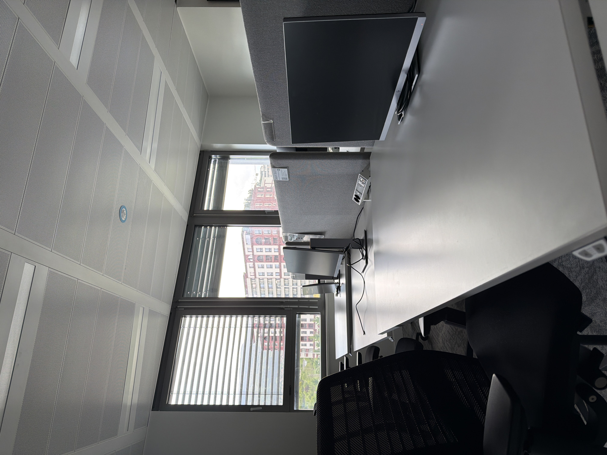 A view from an office in Sofia, Bulgaria. The office space is lined with perforated walls and long windows with blinds. Outside the windows, there are buildings and greenery. Inside, there's a long white desk with computer monitors and chairs. The workspace is partitioned with gray fabric dividers.
