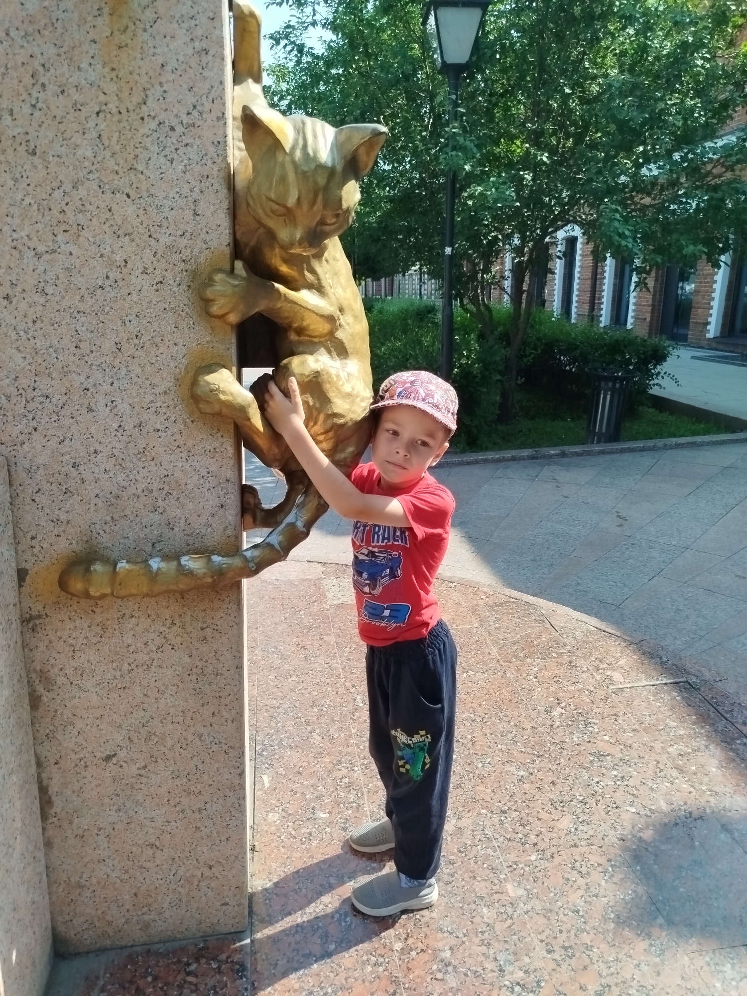 In the city of Tyumen, Russia, a young boy in a red t-shirt, dark pants, a patterned cap, and gray sneakers is standing next to a bronze sculpture of a cat climbing a granite pillar. The boy's right hand is touching the sculpture. The sculpture appears to be part of an outdoor installation. The background shows a paved area with scattered shadows, trees, a lamppost, and a brick building. The sky is visible and appears to be clear.