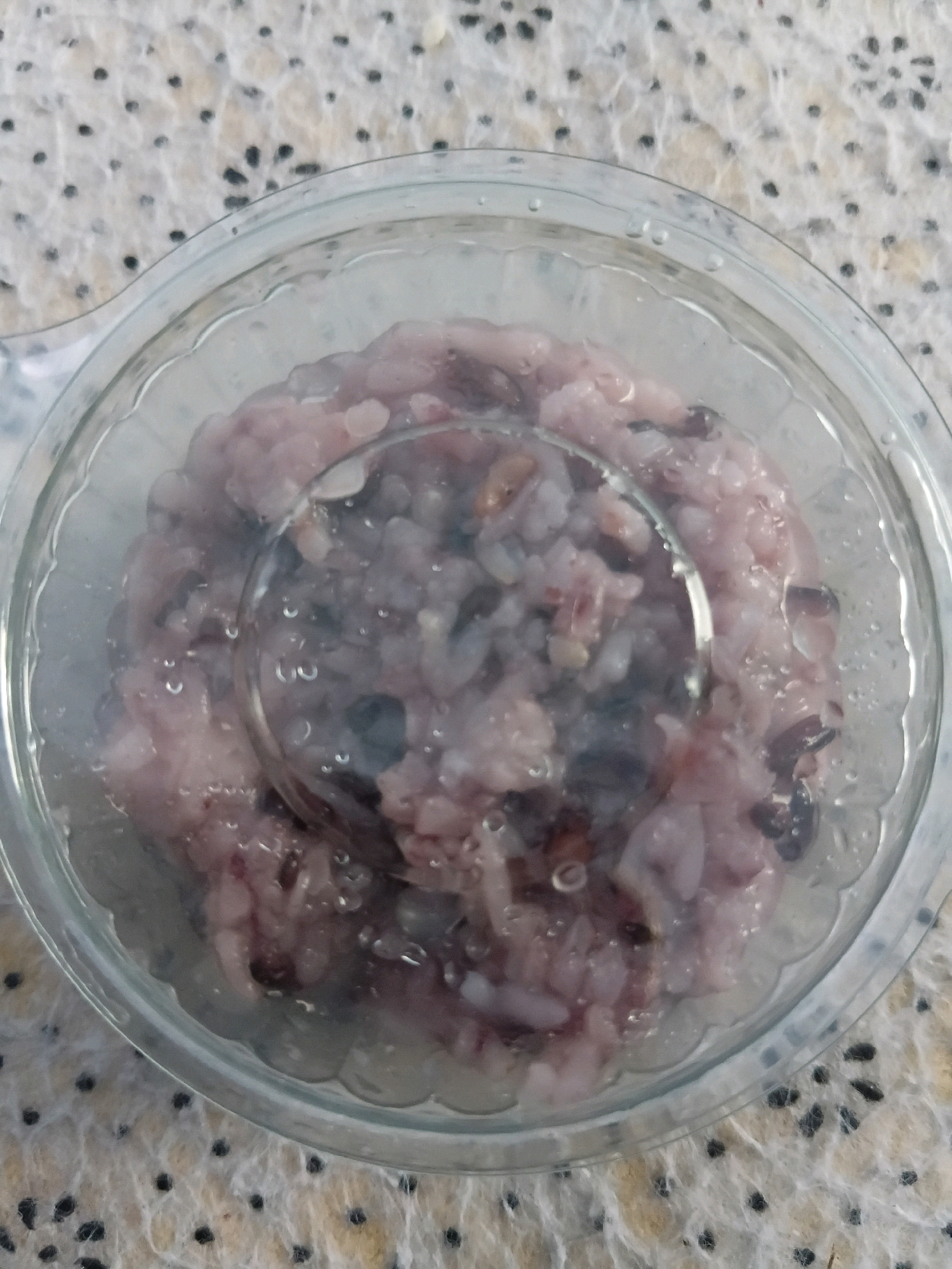 A close-up, overhead shot shows a clear plastic container filled with a pinkish-purple rice pudding. The pudding has visible darker specks throughout, likely from black rice or berries. The container is round with a scalloped edge and is placed on a textured, speckled surface that appears to be a tablecloth or placemat. The lighting is even, and there are some condensation droplets on the inside of the container's lid. The image lacks any specific details that would indicate its location in Tanggul Wetan, Indonesia.