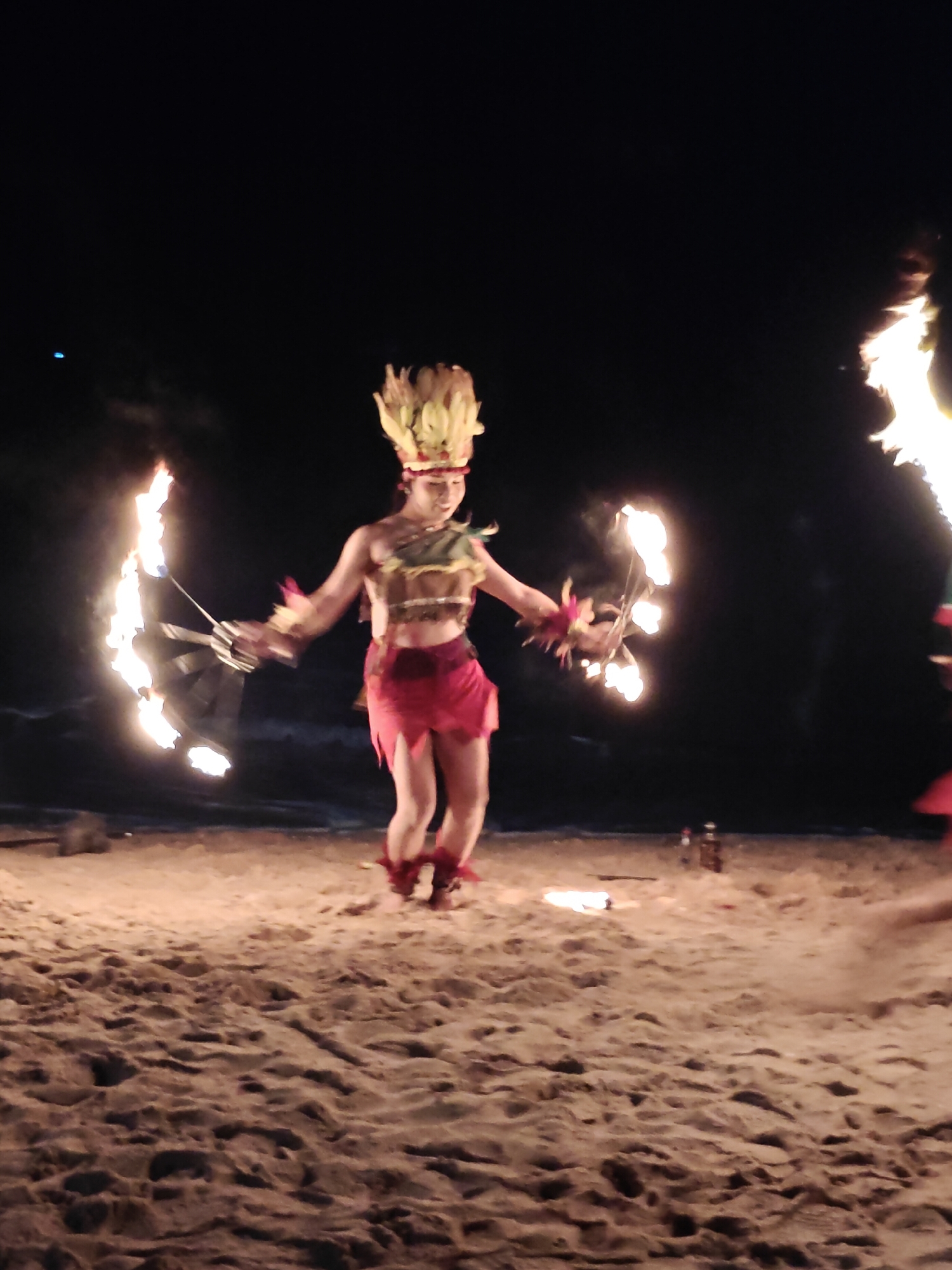 A fire dancer performs on a sandy beach at night, with the ocean visible in the background. The dancer, a person of Asian descent, is wearing a traditional costume consisting of a headdress, a beaded top, and a red skirt. They are holding burning props in each hand, creating a spectacle of fire. The beach is illuminated by the flames, casting a warm glow on the sand. The overall atmosphere is one of a lively cultural performance, likely taking place during a tourist event in Pattaya City, Thailand.