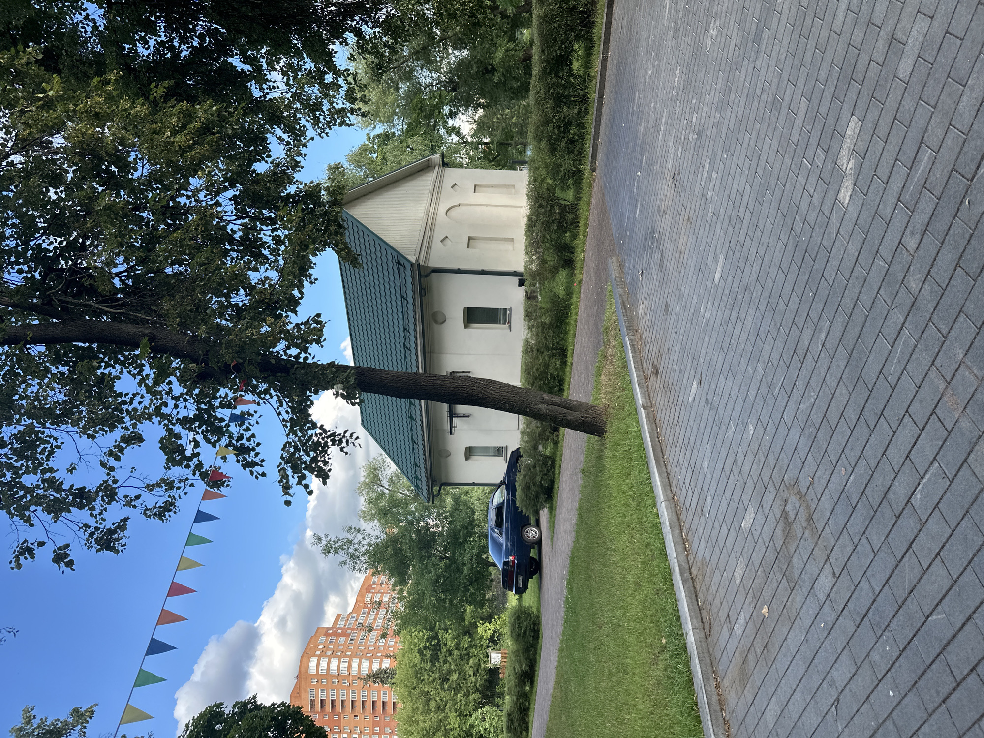 In Kotelniki, Russia, a dark blue sedan is parked on a paved driveway next to a small, white building with a green roof. The building has two arched niches on its facade and a single window on the left. A mature tree with dark green leaves stands in front of the building. The driveway is constructed of gray interlocking pavers, bordered by a strip of green grass and a concrete curb. In the background, a tall, orange residential building with many windows is visible above a line of lush green trees. A string of colorful triangular flags hangs across the sky. The sky is blue with scattered white clouds, and the overall scene appears to be a sunny day in a residential area.