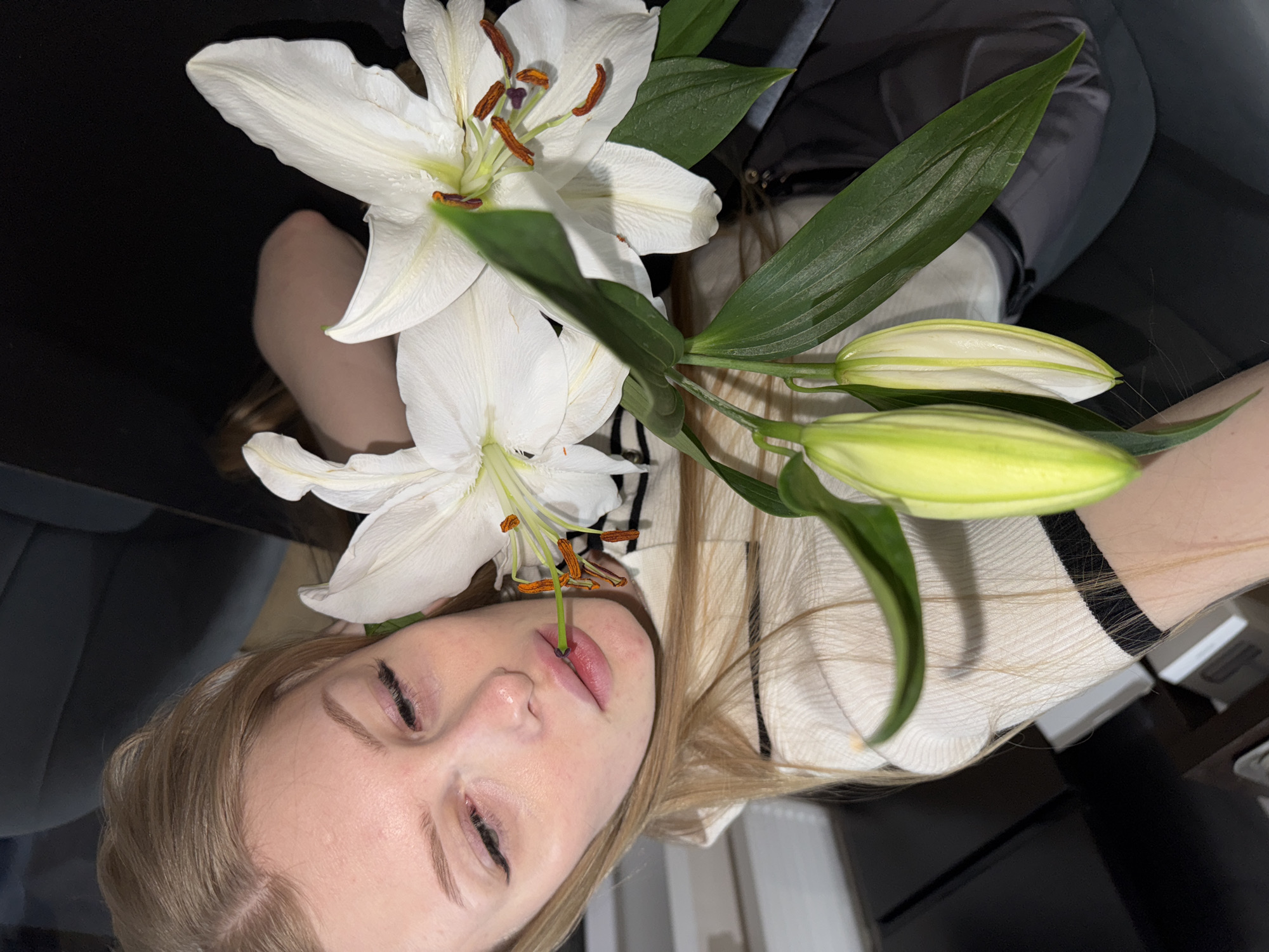 A young, fair-skinned person with closed eyes and blonde hair is positioned beneath a bouquet of white lilies. A single stem of the lily is placed between the person's lips. The person is wearing a light-colored, striped garment. The background consists of a dark-colored, upholstered chair or sofa, as well as other furniture and objects. There is no indication of the image being taken in Perm, Russia.