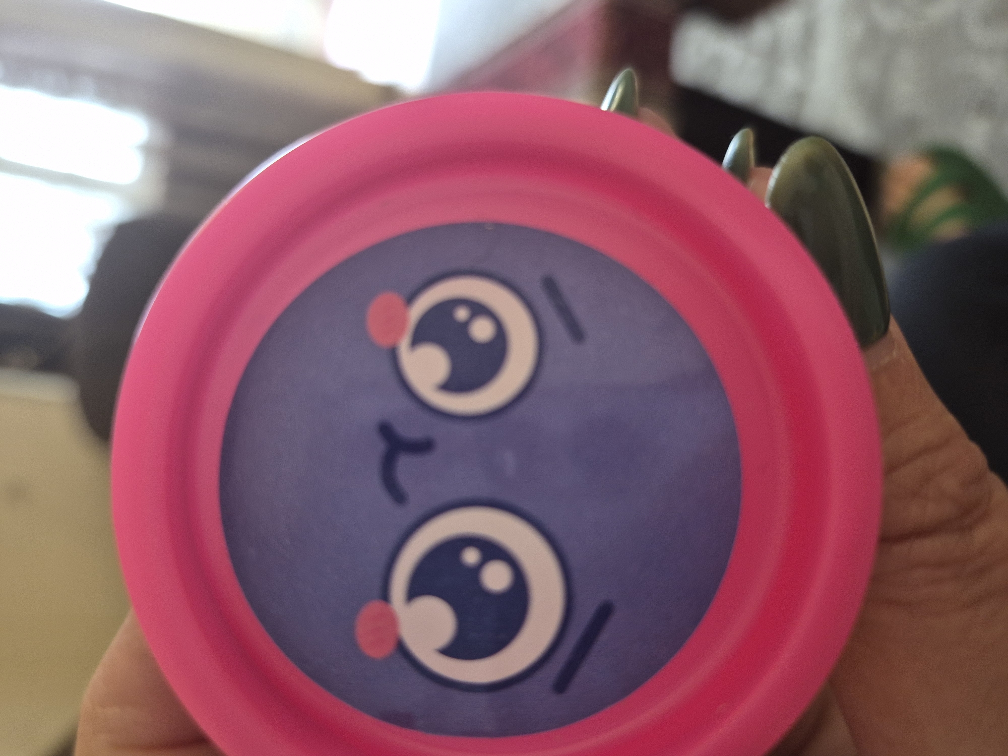 A close-up, slightly blurry photograph features a circular object with a cartoon face, held by a person's hand. The object has a bright pink outer rim and a purple center. On the purple surface, a stylized cartoon face is depicted, oriented sideways. It consists of two large, wide-open eyes with white sclera, blue irises, and white highlights, giving a shiny or surprised look. Small, round pink blush marks are placed below each eye. Simple, short black lines represent eyebrows and a tiny, slightly curved line suggests a mouth.
The object is held from the lower right side by what appears to be fingers. The person's fingernails are prominent, long, and oval-shaped, painted a dark, olive-green color. The skin tone of the hand is visible.
The background is heavily blurred, suggesting an indoor setting with bright, diffused light, possibly from a window or artificial lighting. Faint shapes and colors in the background might indicate furniture or room elements. There is no visible text in the image. The overall impression is a casual, handheld shot of a playful object.