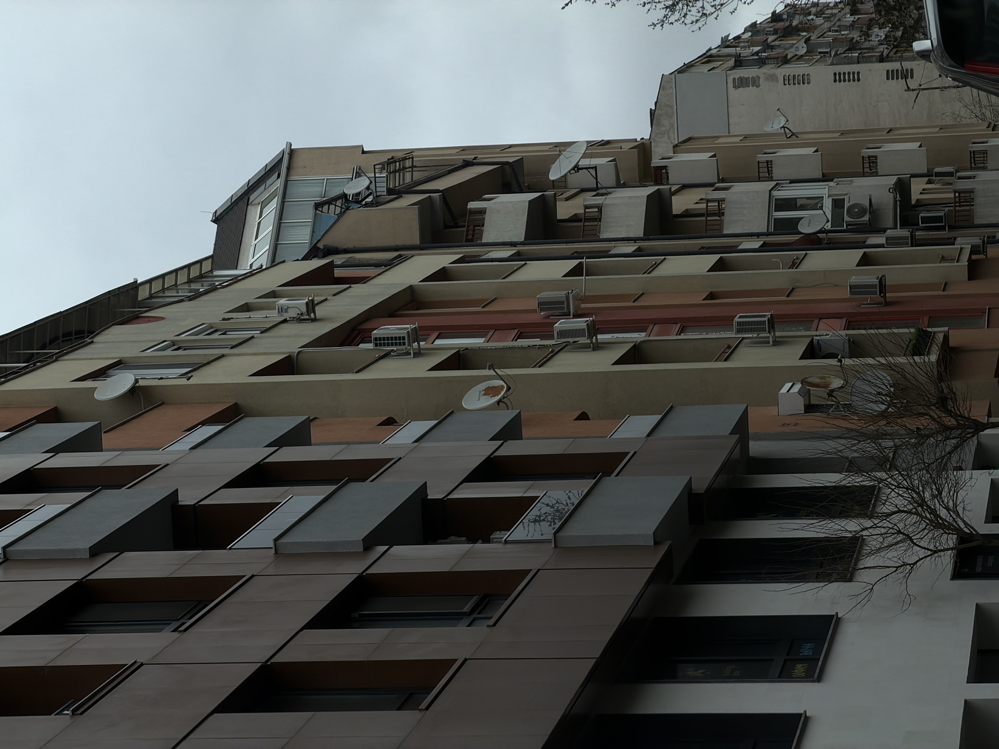 The image captures a low-angle perspective of a unique residential building in Sofia, Bulgaria. The building features a stepped architectural design with varying facade treatments on each level. The lower floors are characterized by dark brown panels creating a pattern of recessed windows. Mid-levels feature a lighter color scheme, incorporating windows and balconies. Satellite dishes and air conditioning units are scattered across the mid and upper levels. The sky is overcast. A bare tree branch is visible in the foreground on the right side, and a vehicle partially visible in the upper right corner.