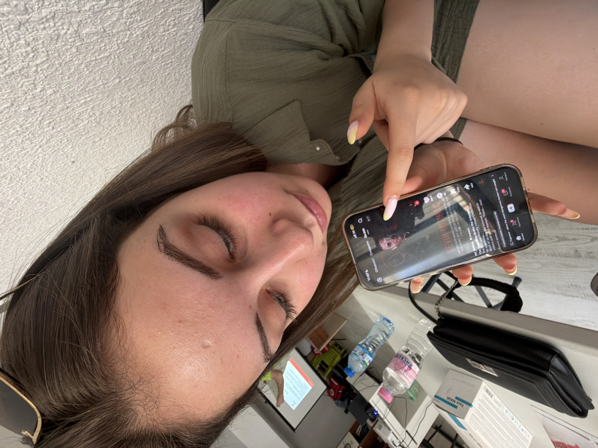 A young woman with brown hair and fair skin is looking down at her smartphone, which she is holding in her right hand. Her left index finger is pointing at the phone screen. She is wearing a green collared shirt. In the background, a projector screen with a presentation is visible, along with two bottles of water on a table, a black purse, and a box labeled "Face Mask." The scene appears to be indoors, possibly an office or classroom setting. There is no visible indication of the city of Burgas, Bulgaria.