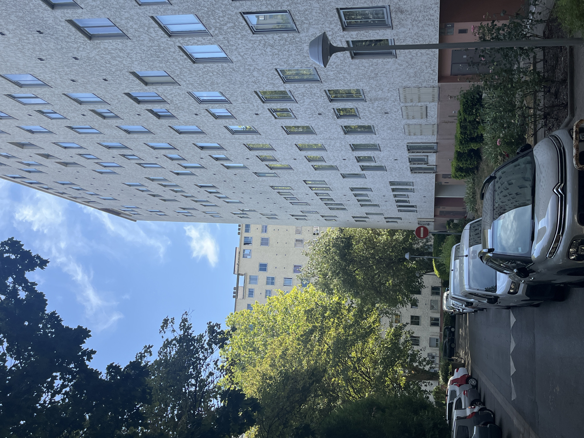 The image depicts an outdoor scene in Fresnes, France, with a focus on residential buildings and parked cars. In the foreground and to the right, a light-colored, modern apartment building with numerous windows dominates the frame. The building's facade is textured and off-white. A street lamp stands in front of it. To the left, a line of parked cars is visible on a paved street. The cars vary in color and make, including a silver Citroen in the immediate foreground.
Behind the parked cars, a large, leafy green tree casts shadows on the street and the adjacent building. Further in the background, another residential building is visible, appearing lighter in color and with multiple windows. The sky above is blue with scattered white clouds, and the sunlight suggests it is daytime. The overall environment suggests a residential area, likely a housing complex. There are no people or animals visible in the image. The scene appears calm and orderly, with an emphasis on architecture and infrastructure.