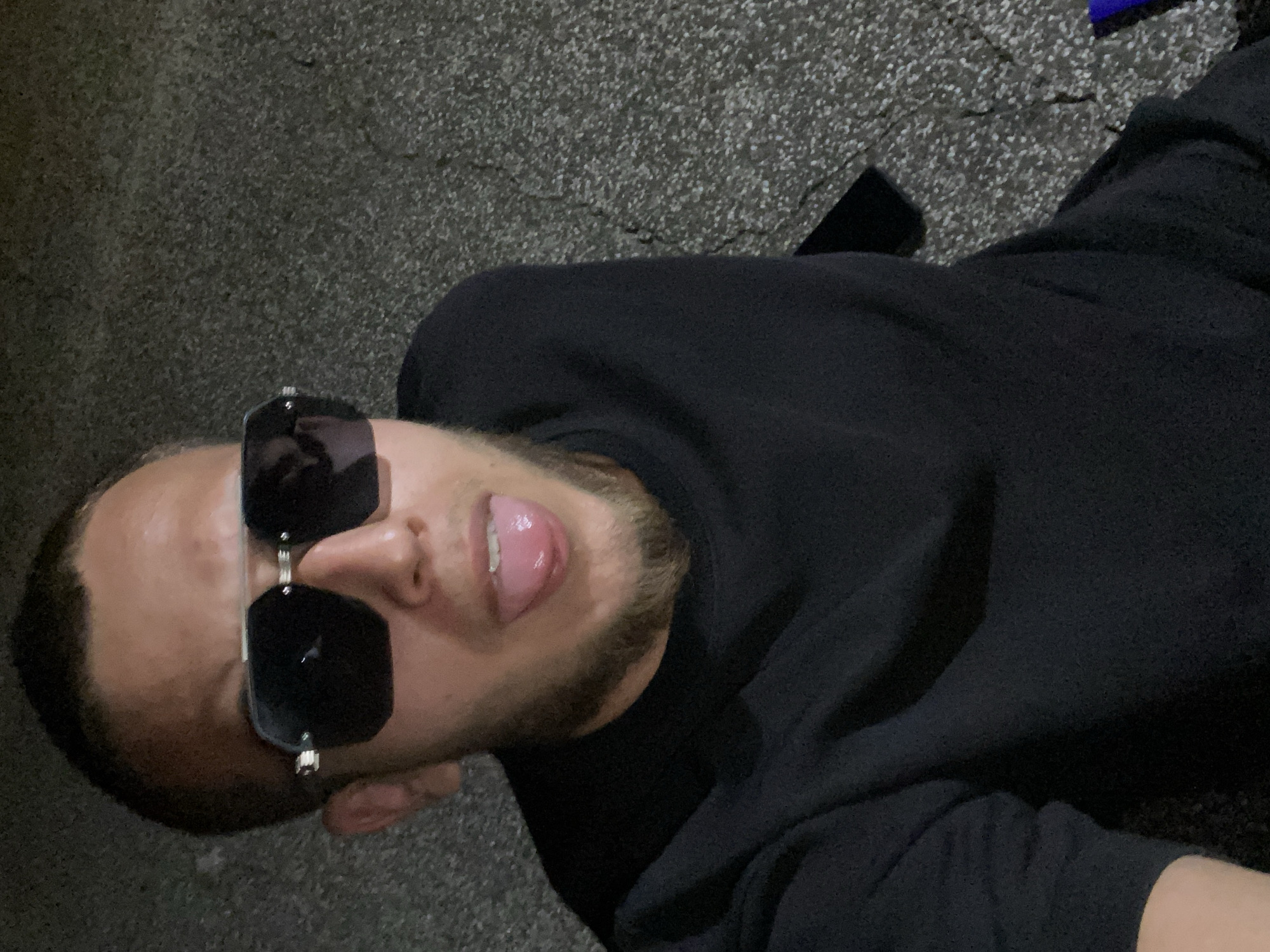 A man wearing a black shirt and sunglasses sticks his tongue out at the camera. He has a short beard and is outdoors with a gritty, asphalt-like ground in the background. The image is a close-up, focusing on his face and upper body. There is no indication of the city of Hirevo, Bulgaria, or any specific location details in the visual information provided.