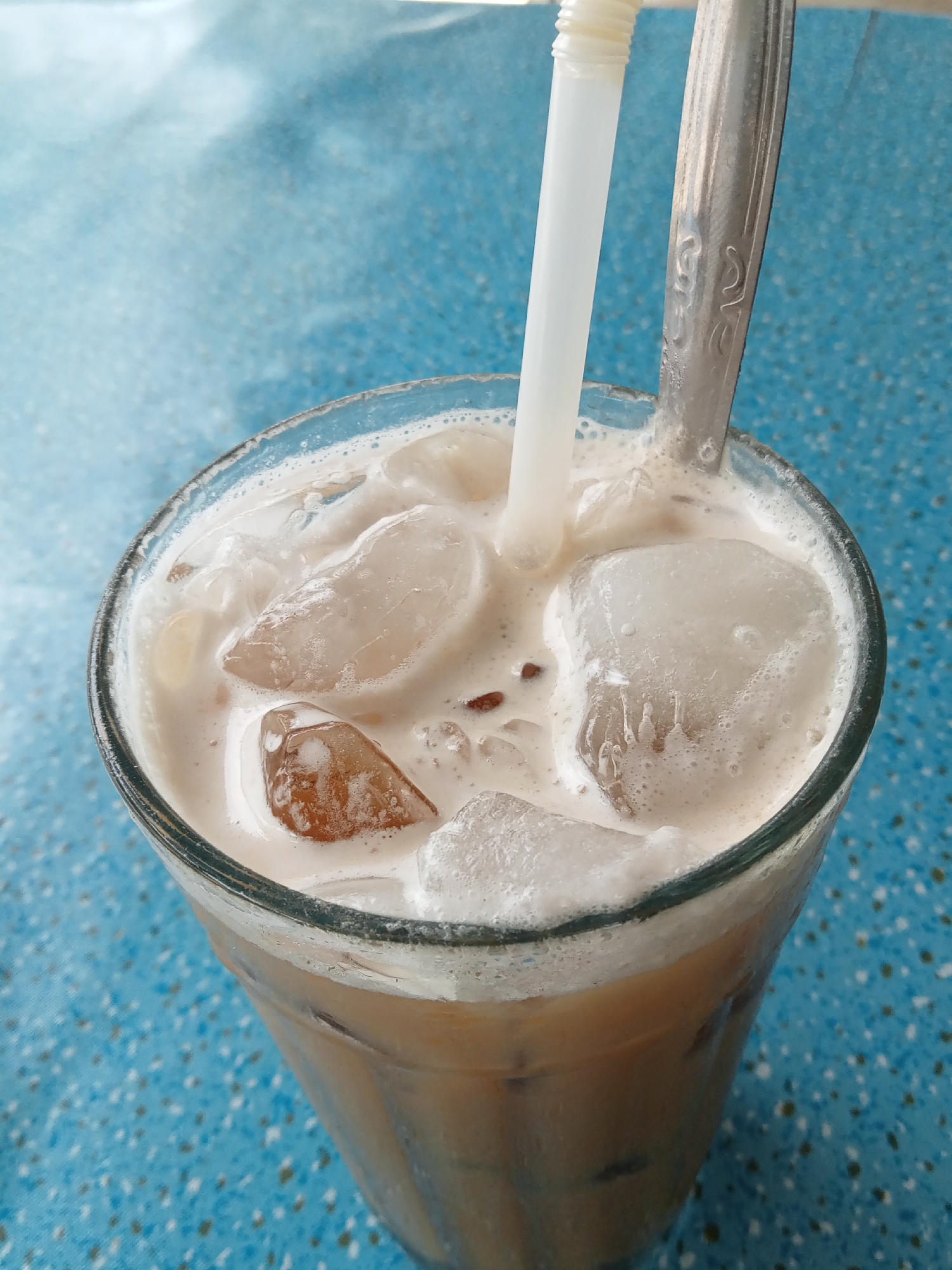 A close-up, high-angle shot shows a glass of iced coffee on a blue, textured surface. The glass is filled with a light brown liquid, ice cubes, and a foamy layer on top. A white plastic straw and a silver spoon are inserted into the drink. The blue surface appears to be a table. The scene is indoors, likely in a cafe or restaurant. The location is specified as Matangsijuek Timur, Indonesia. There are no people visible in the image.
