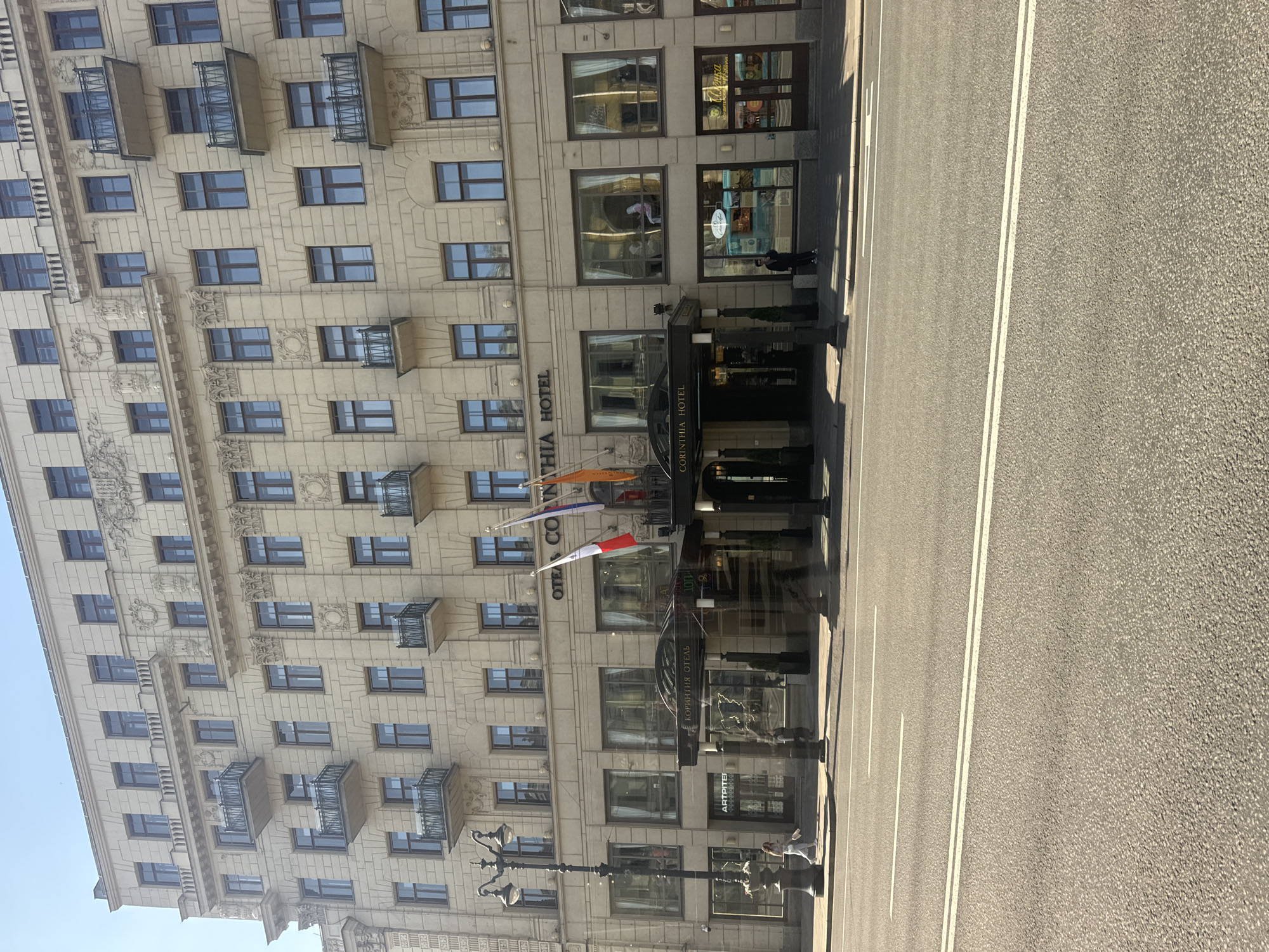The image shows a street view of the Corinthia Hotel in Saint Petersburg, Russia. The hotel is a large, ornate building with multiple floors and many windows. The facade is decorated with intricate carvings and balconies. In front of the hotel, there is an entrance canopy with the hotel's name written above it. Flags are visible near the entrance, including the French flag. A street lamp stands on the sidewalk to the left of the entrance. A woman in light-colored clothing is walking on the sidewalk in front of the hotel. Across the street, a paved road with white lane markings is visible. The sky is clear and blue.