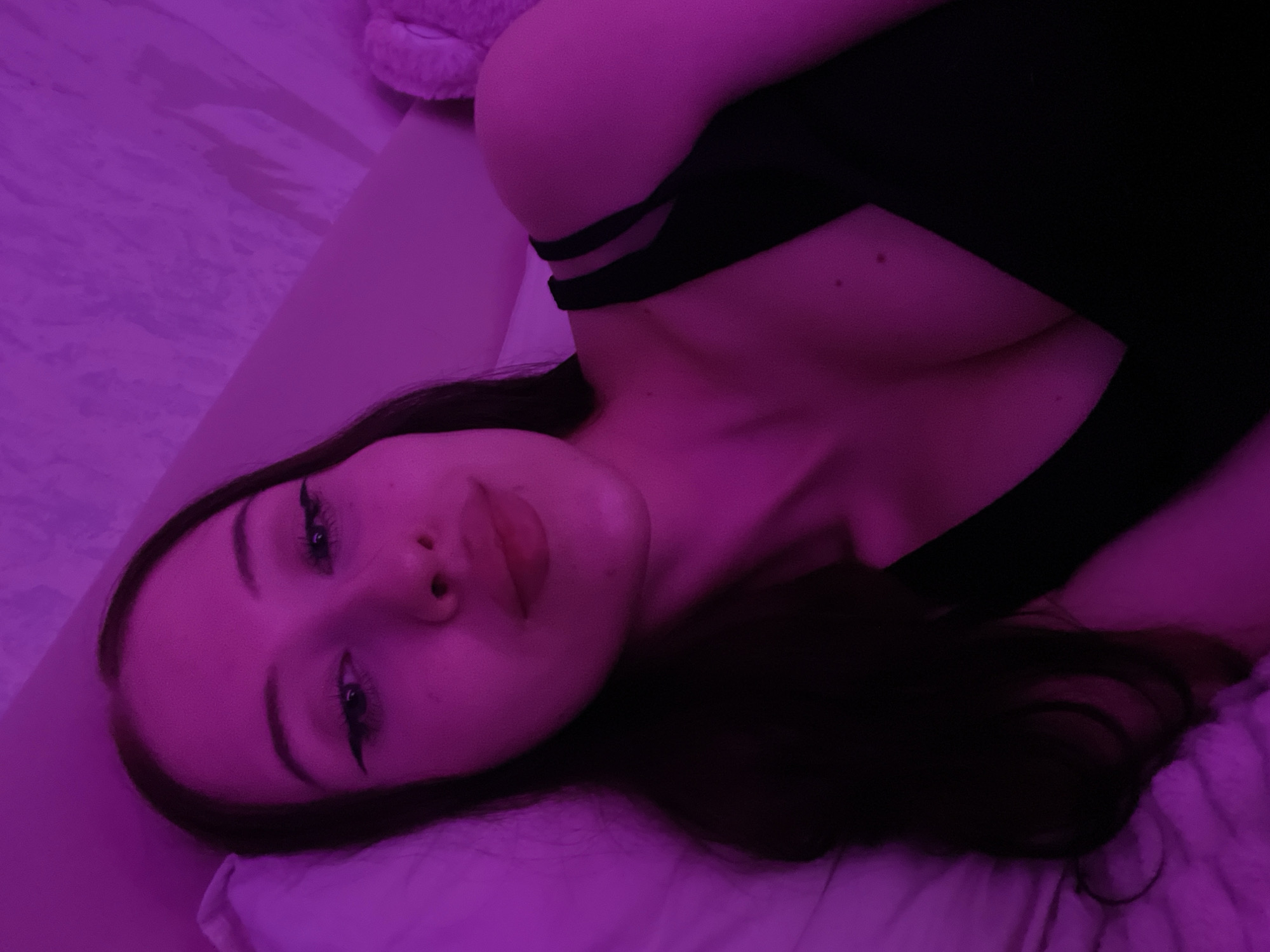 The image depicts a young woman lying down in a dimly lit room with purple lighting. She has long, dark hair and is wearing a black top. Her head is tilted, and she is looking towards the camera with winged eyeliner. The background appears to be a bed with a pink-toned textured surface. The context of Votkinsk, Russia, cannot be determined from the visual information presented in the image.