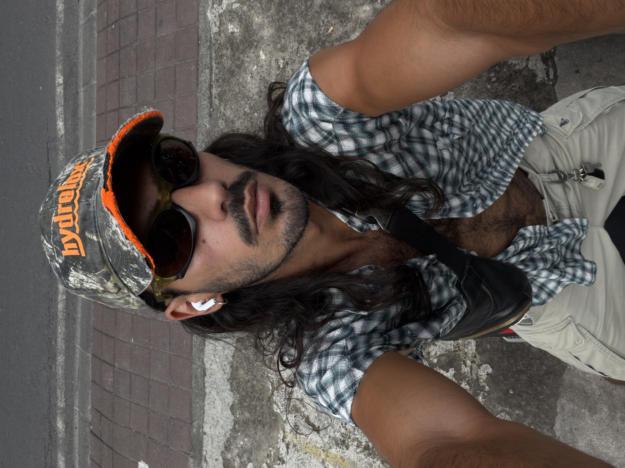 An overhead selfie captures a man lying or reclining outdoors in North Kuta, Indonesia. The man has long dark hair, a beard, and a mustache. He is wearing a camouflage-patterned baseball cap with an orange brim, featuring the word "hydrelox" in orange text. Dark sunglasses cover his eyes, and a white earbud is visible in his left ear.
He is dressed in an unbuttoned green and white plaid long-sleeved shirt, revealing his chest hair, and light-colored (khaki or beige) pants or shorts. A black fanny pack or crossbody bag, with a subtle red and green stripe detail near its zipper, is worn across his chest. A set of keys on a keychain hangs from his pants near his hip. His right arm is extended upwards, indicating he is holding the camera or phone.
The setting appears to be a paved outdoor area, possibly a street or pathway, with sections of dark grey pavement and lighter textured ground visible. The scene is brightly lit, suggesting daytime. The man's posture appears relaxed.