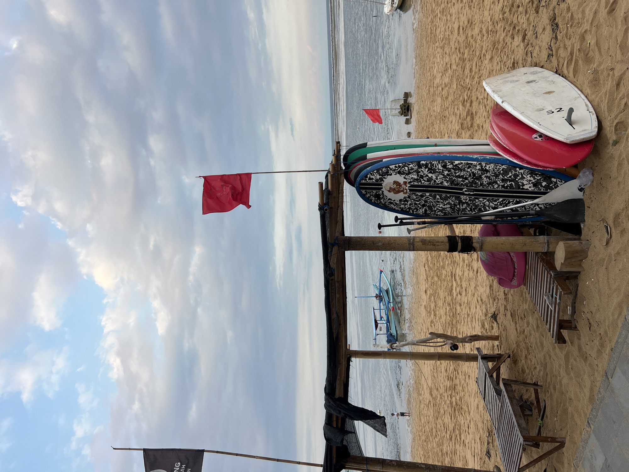 The image depicts a beach scene in Denpasar, Indonesia, under a cloudy sky. In the foreground, on the sandy beach, are several surfboards and paddleboards leaning against a wooden structure. Two wooden lounge chairs are also visible, along with a pink beanbag chair. A small blue and white boat is anchored in the shallow water in the middle ground. Two red flags are planted in the sand, one on a pole near the surfboards and another further out in the water. A person is standing on the beach near the water's edge, appearing to be a child. The environment suggests a recreational area, possibly a surf school or rental shop, given the presence of surf and paddleboards. The lighting indicates it might be early morning or late afternoon.