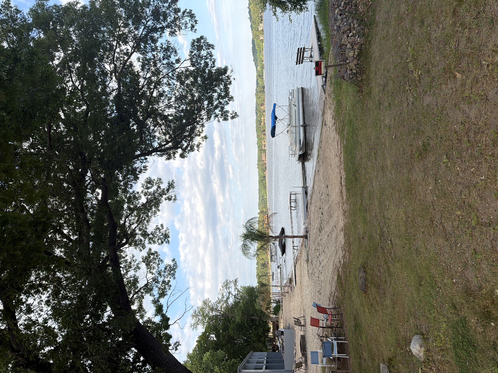 This image captures a serene lakeside scene at Lake Delton, United States of America. A large, verdant tree with dense foliage occupies the left side of the frame, its branches reaching upwards towards a partially cloudy sky. The foreground features a sandy beach area on the left, transitioning into a grassy patch on the right.
At the water's edge, a white pontoon boat with a blue canopy is partially beached or docked, indicating a recreational setting. Further along the sandy shore, a lone palm tree adds to the relaxed, beach-like atmosphere. Scattered across the sandy and grassy areas are several outdoor chairs in red, blue, and grey, suggesting a leisure or gathering spot.
Near the boat and water, a wooden bench is visible, and next to it, a red and white sign prominently displays the text "NO TRESPASSING." The shoreline to the right of the sign is reinforced with a mound of rocks. The calm, blue water of the lake stretches into the background, where distant tree-covered hills line the horizon under a bright sky with scattered white clouds. The scene appears to be set during daytime, likely late morning or early afternoon, conveying a peaceful and inviting ambiance, though no people are present.