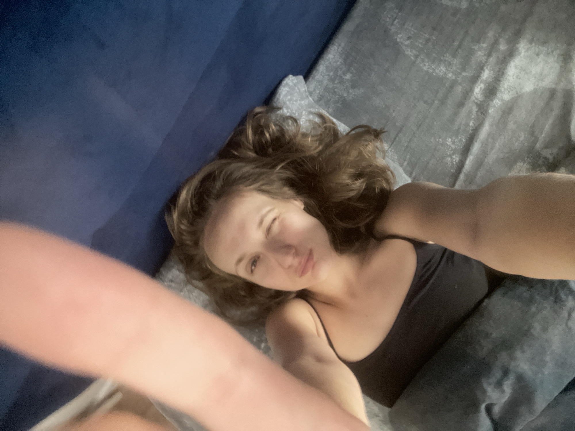 A young woman with light brown hair lies on a bed, looking at the camera and winking. She is wearing a black tank top and is propped up by pillows. The bed has gray sheets and a dark blue headboard. The lighting suggests it is nighttime or indoors with dim lighting. The image appears to be a selfie taken by the woman. The location is specified as Podshivalovo, Russia.