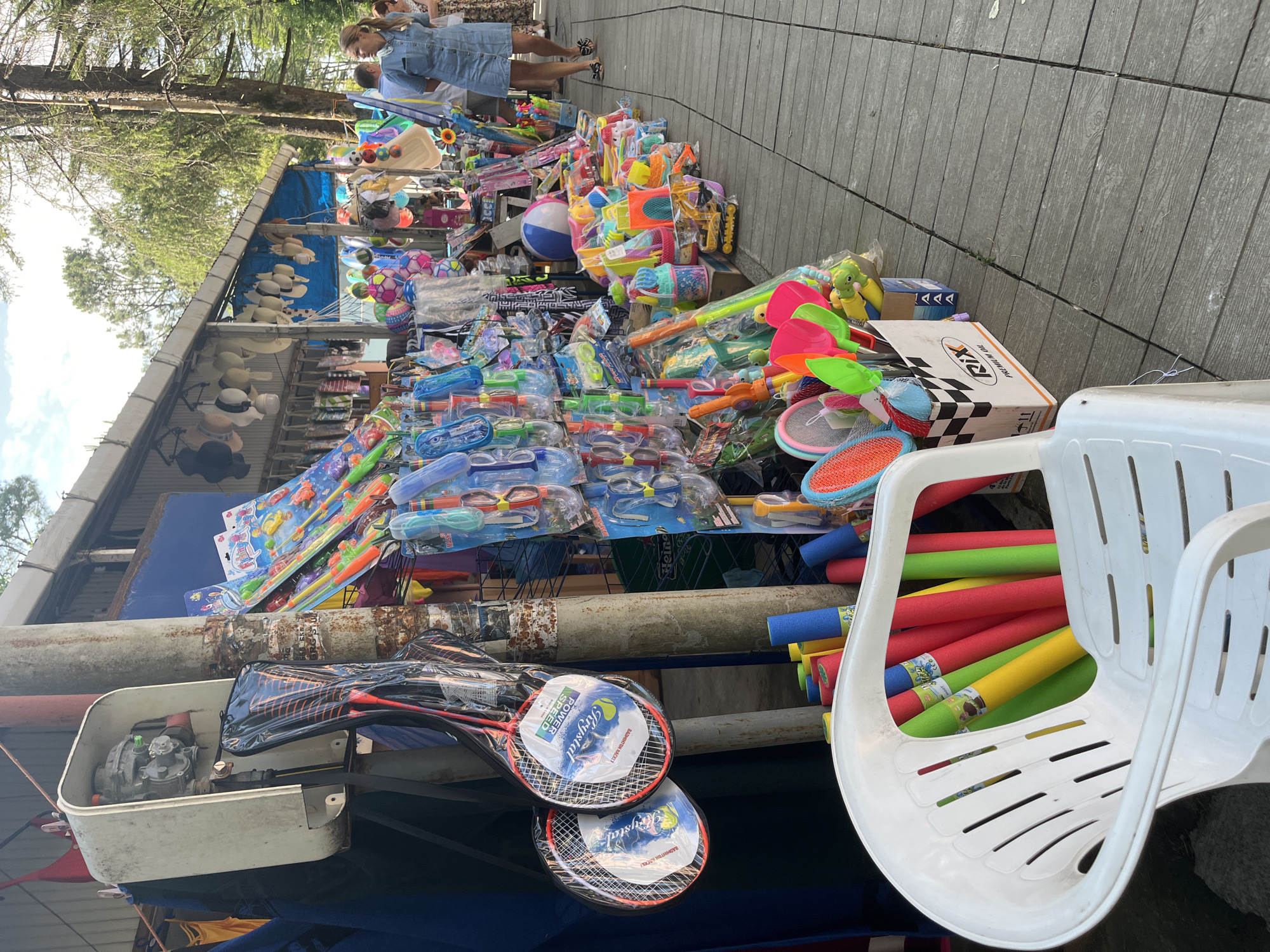 A street market stall in Ureki, Georgia, displays a variety of beach and water toys. The stall is set up outdoors on a wooden boardwalk, with a covered structure in the background that houses more merchandise. In the foreground, a white plastic chair is filled with colorful foam pool noodles in red, yellow, and green. To the right of the chair, a cardboard box overflows with brightly colored plastic shovels and rakes, along with other toys like a small yellow toy construction vehicle. Further down the stall, an assortment of water guns, goggles, and snorkels are neatly displayed on racks. Beach balls, including one large blue and white striped ball, are also visible. A woman in a denim dress is seen walking away from the camera, and another woman with blonde hair is visible in the background, suggesting a public area with pedestrian traffic. The overall impression is of a lively summer market catering to vacationers.