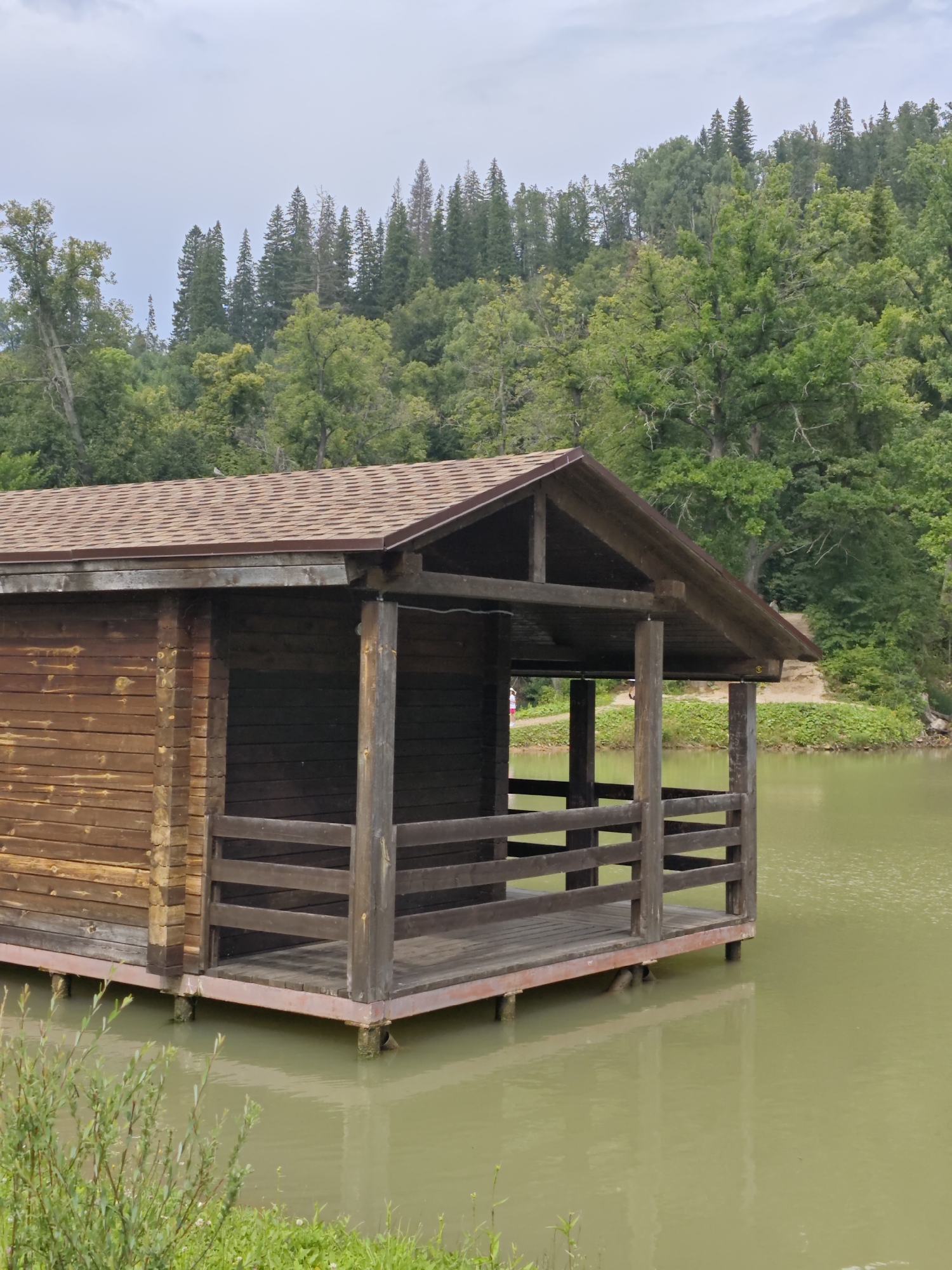 A wooden boathouse floats on a tranquil, murky body of water. The structure is made of horizontal wooden planks and features a covered porch with wooden railings. Behind the boathouse, a dense forest of green trees rises up a hill, under a cloudy sky. In the foreground, lush green vegetation grows along the water's edge. The scene appears to be located in or near the city of Krasnyy Klyuch, Russia.